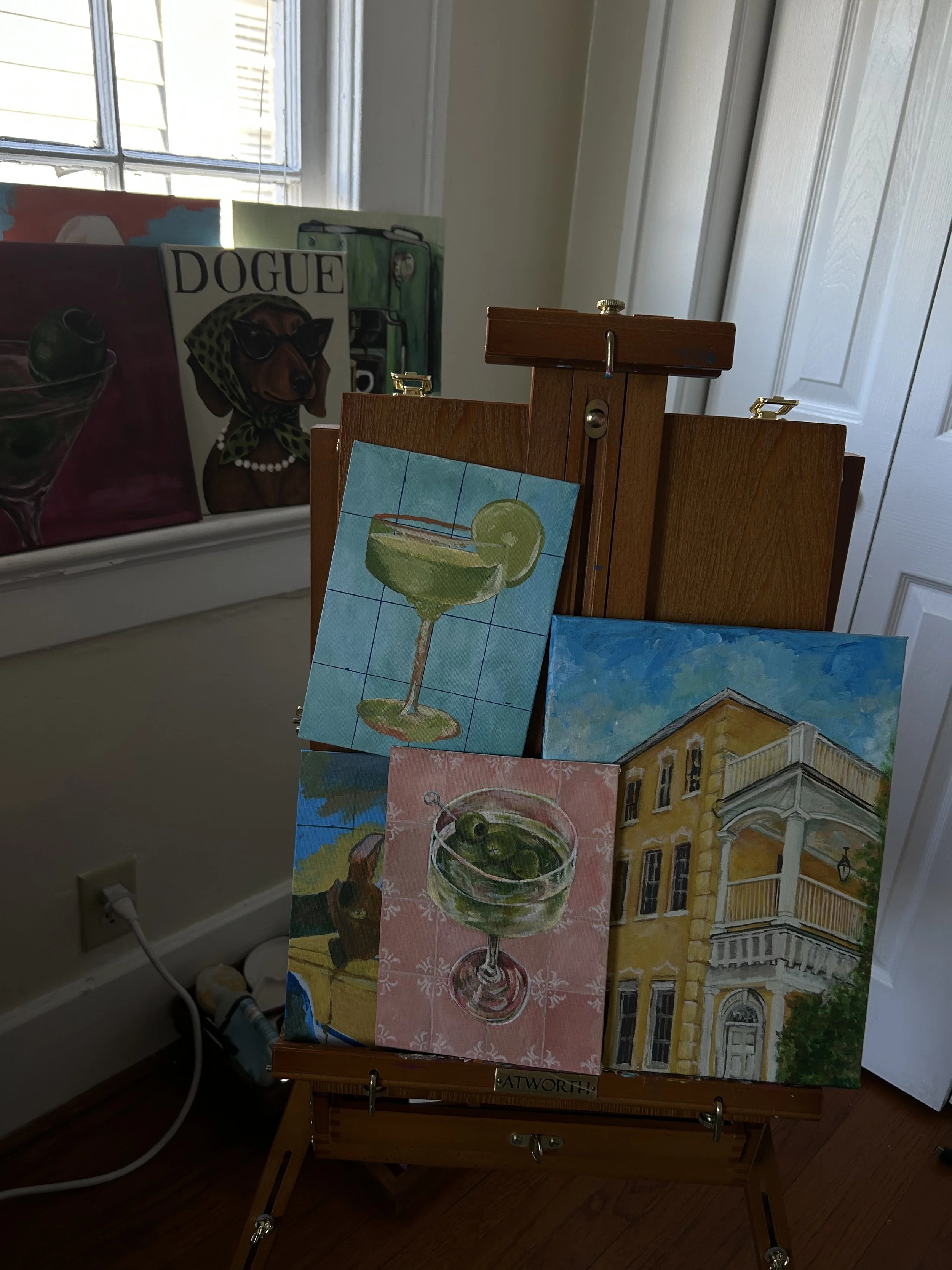 Several paintings on a wooden easel include a colorful house, a green cocktail, and an outdoor scene with a building.