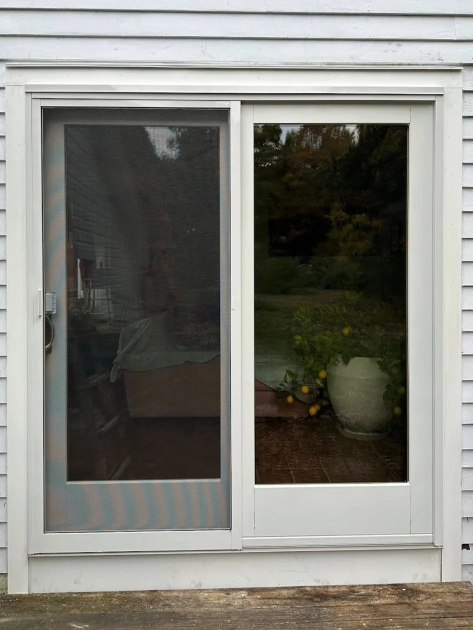 A sliding glass door with a screen on the left and a clear glass panel on the right. Outside, there's a potted lemon tree with yellow lemons, and a reflection of trees and sky in the glass.