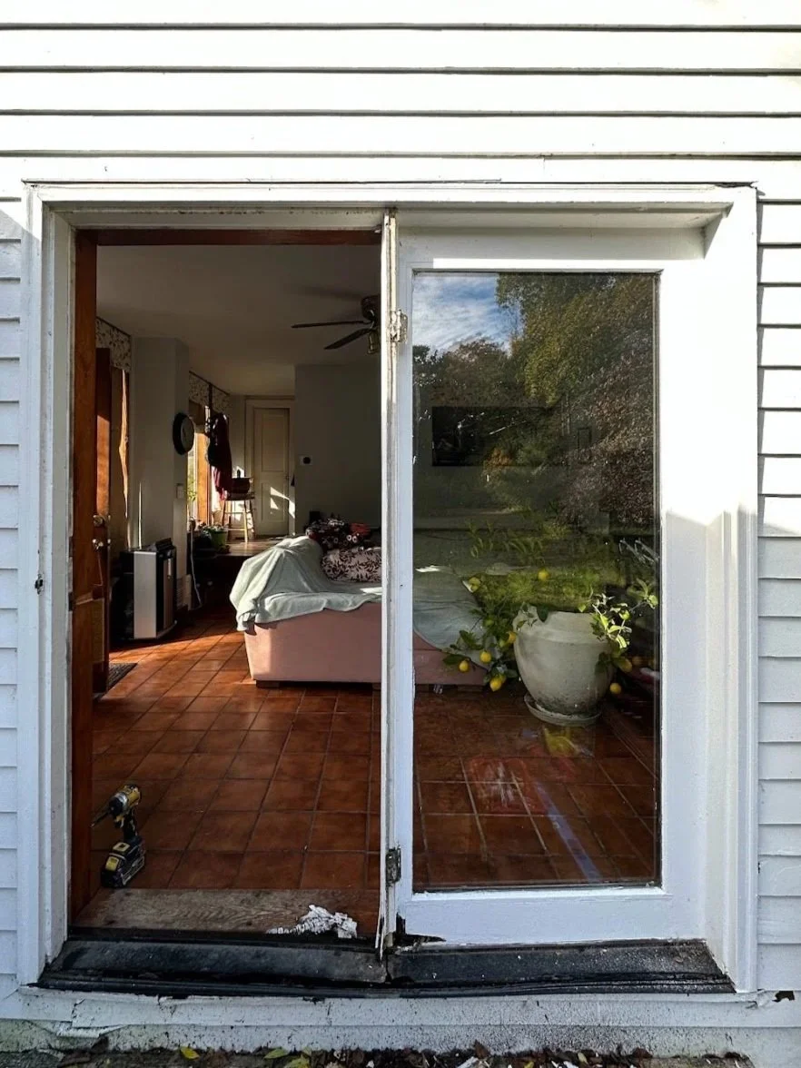 View through a sliding glass door showing an interior living room with tile flooring, a sofa covered with a cloth, a ceiling fan, and a large potted lemon tree with yellow lemons outside on the patio.