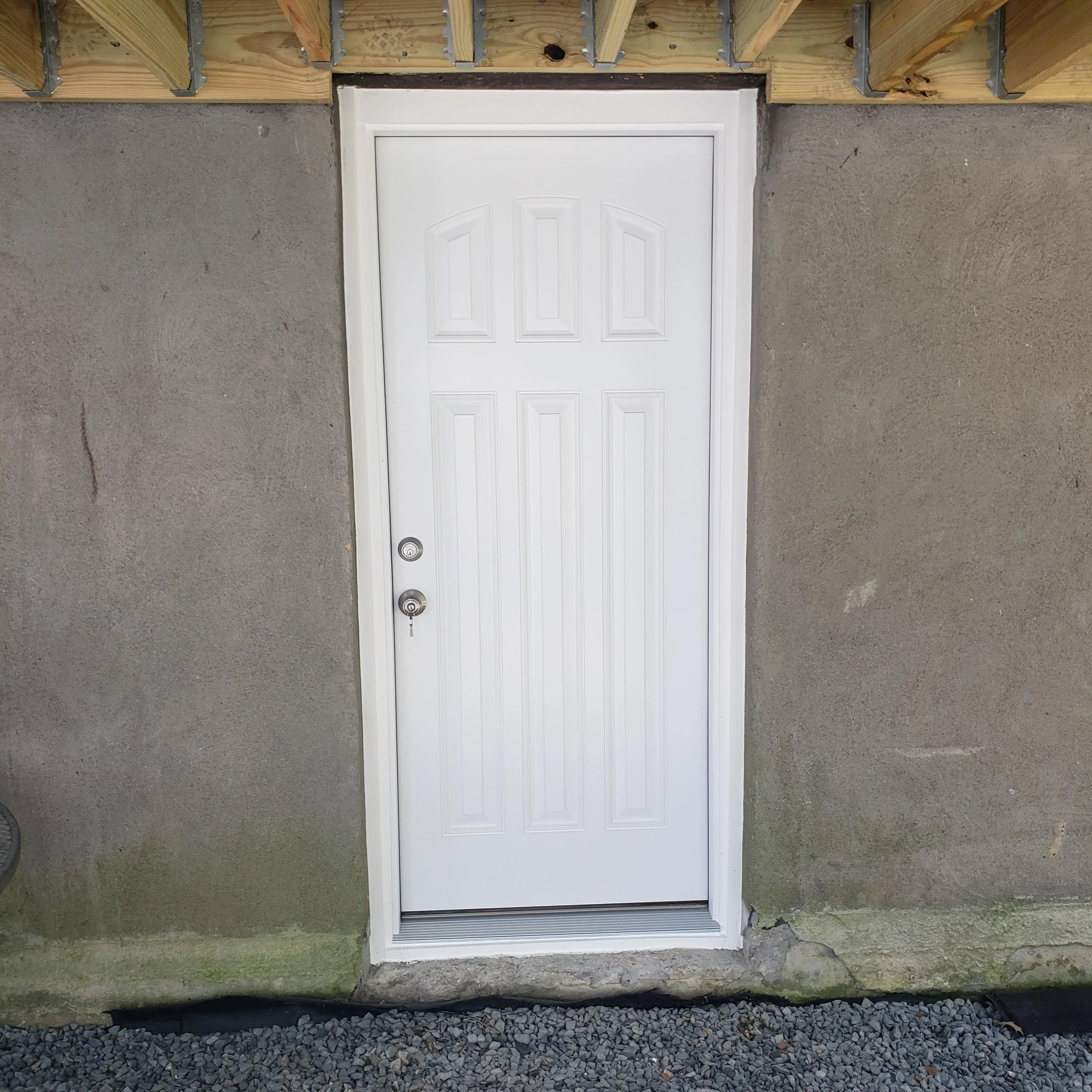 White door with panels and a doorknob, set in a concrete wall with gravel at the base, under construction with exposed wood framework above.