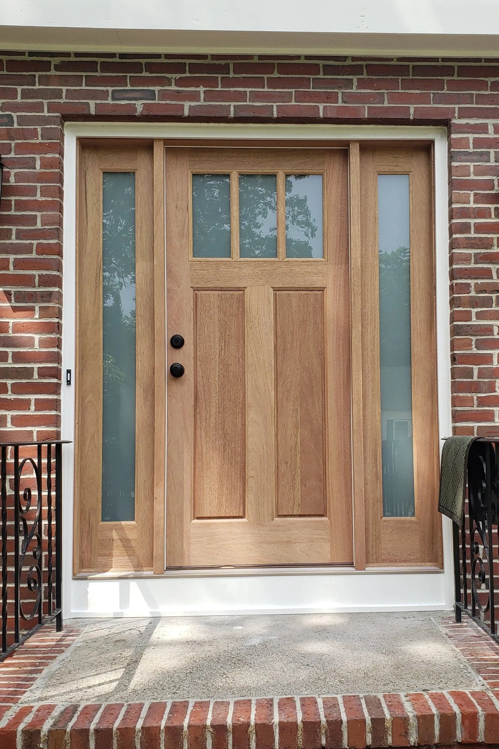 Front door with a wooden finish, flanked by two narrow glass panels, set in a brick house with a small concrete porch, a black metal railing on each side, and a doormat on the porch.