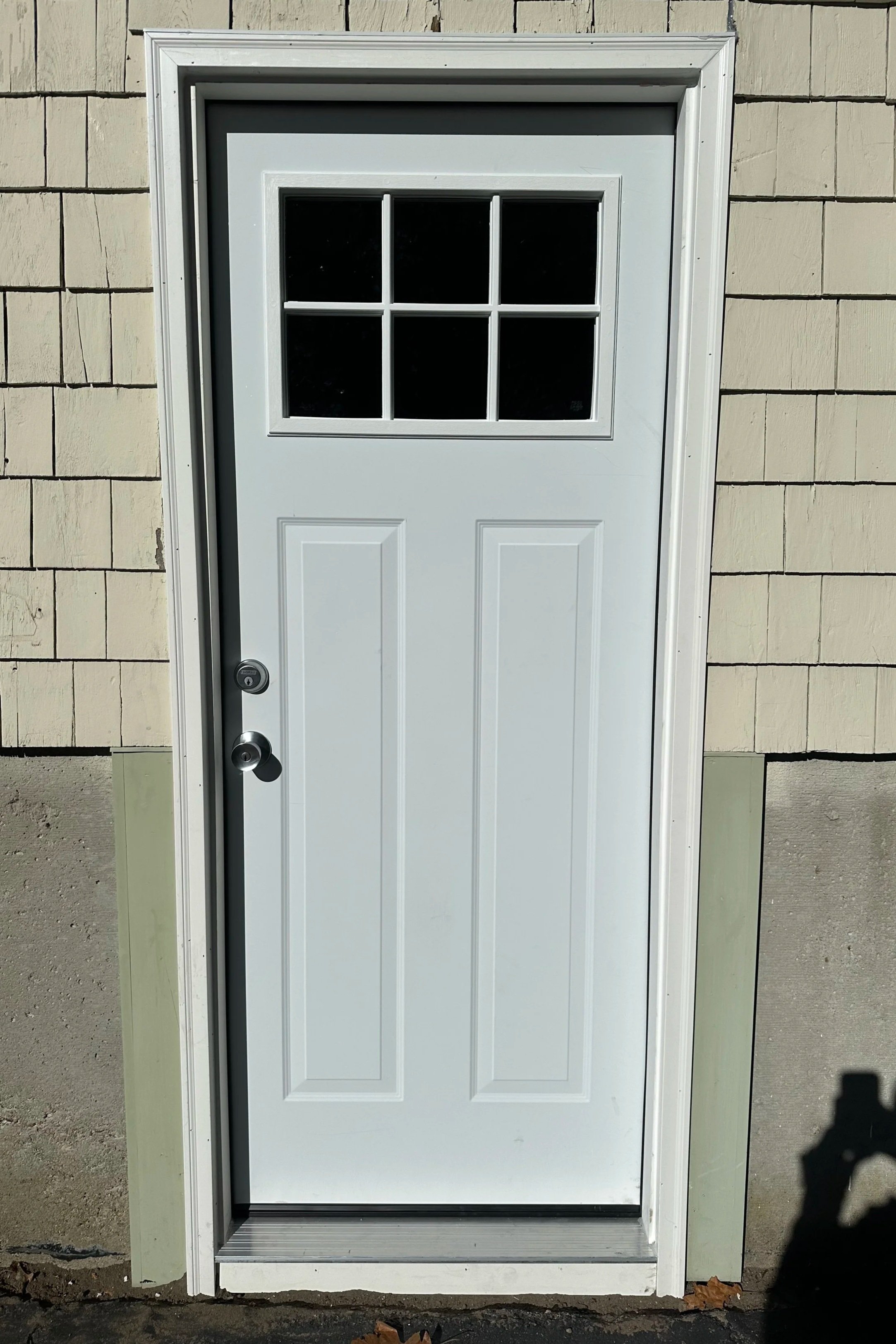 White exterior door with six-pane window at the top, installed in a building with beige brick siding, shadow of a person taking the photo is visible on the lower right side.