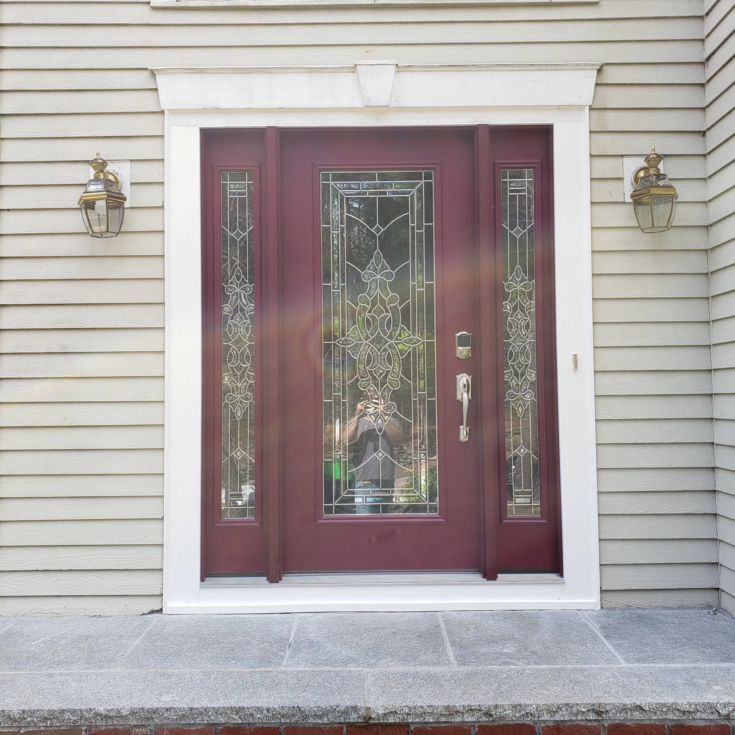 Front door with decorative glass panels, maroon frame, and beige siding with horizontal planks. Two brass outdoor lanterns are mounted on the wall beside the door.