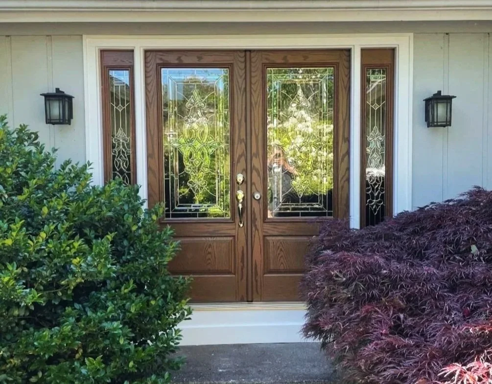 Front door with decorative glass panels, flanked by two wall-mounted lanterns, surrounded by green and purple bushes.