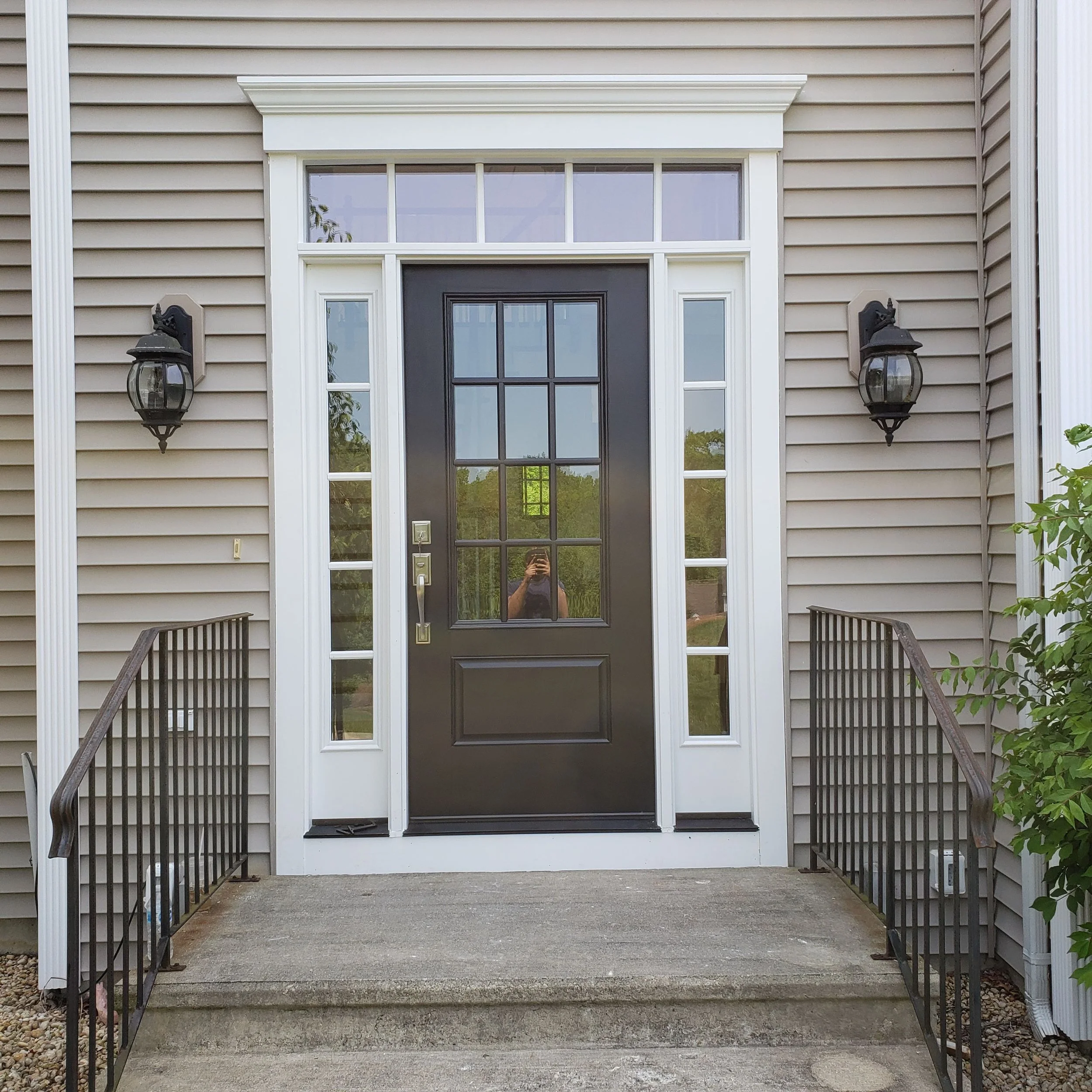 Front view of a house entrance with a black door, surrounded by white trim, with two black wall mounted lanterns on each side, a concrete porch with stairs, and beige vinyl siding.