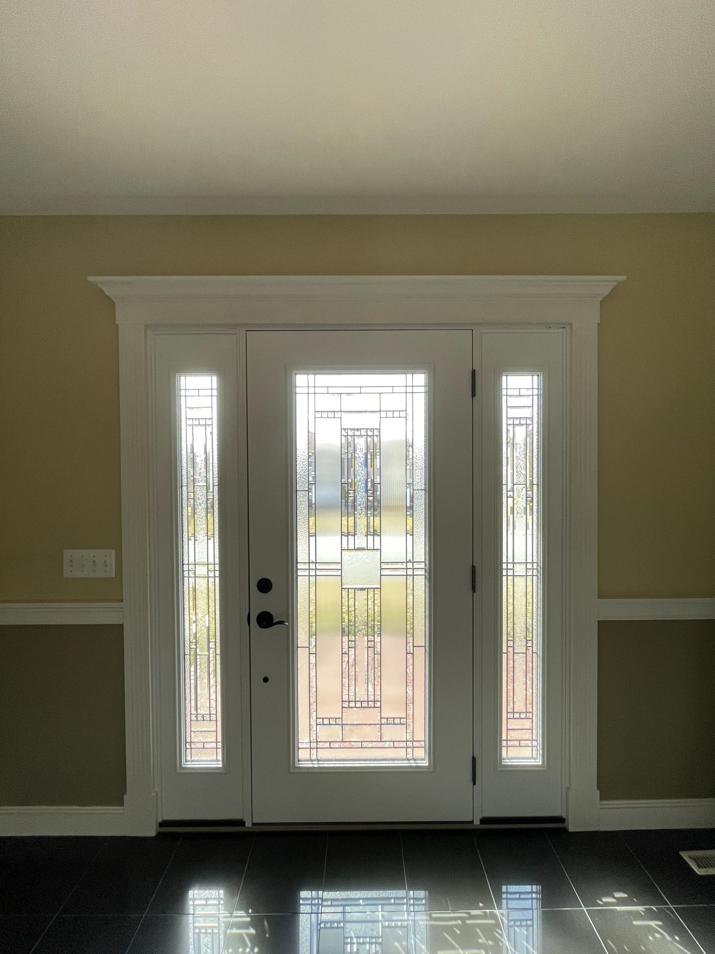 A white front door with decorative glass panels and sidelights, set in a yellow wall with white trim, and a dark tiled floor reflecting light.
