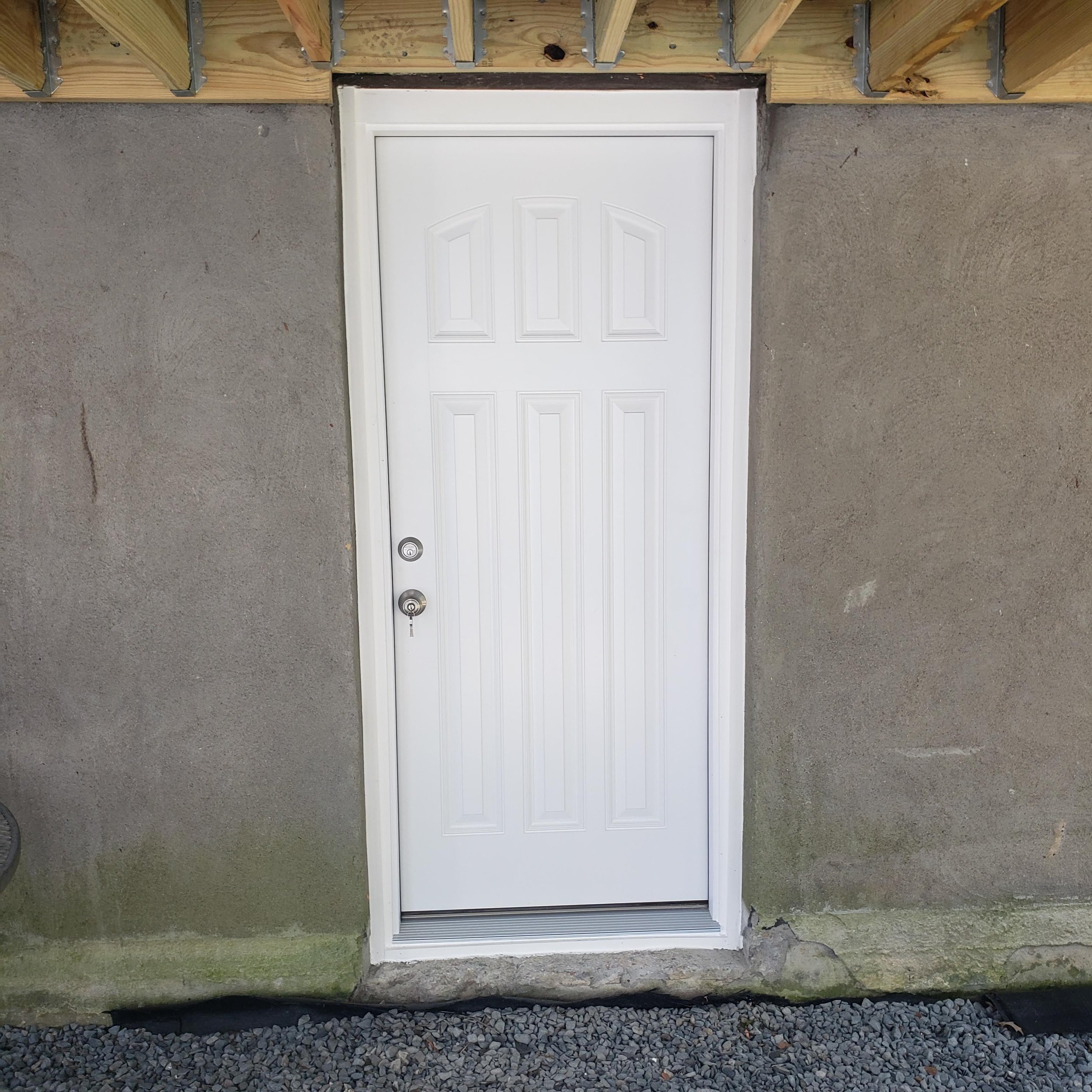 Front view of a white residential door with a security lock, set in a wall under construction with visible framing, concrete, and gravel at the bottom.