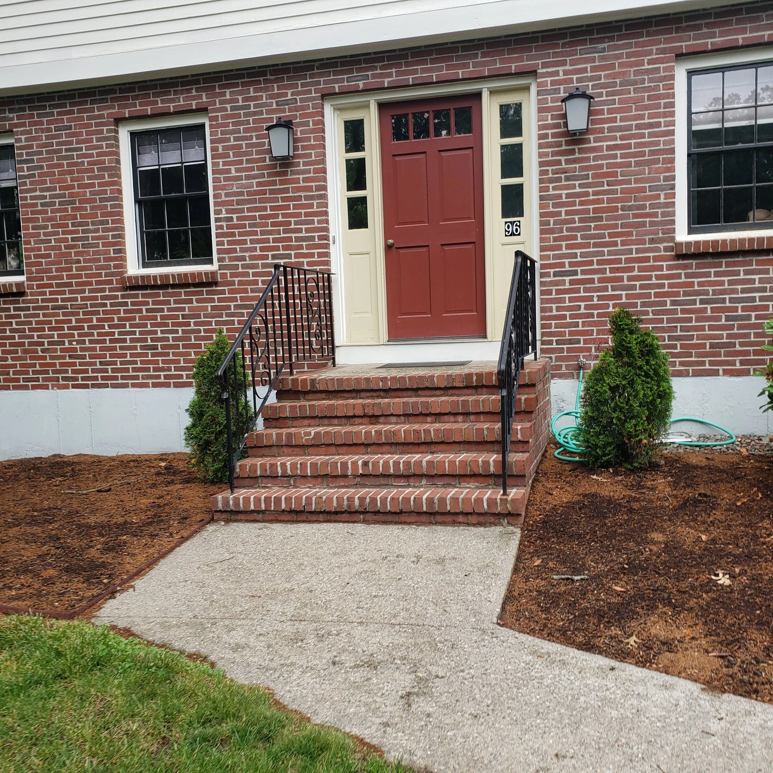 Front entrance of a brick house with a red door, black railings, two small bushes, and a garden hose on the ground.