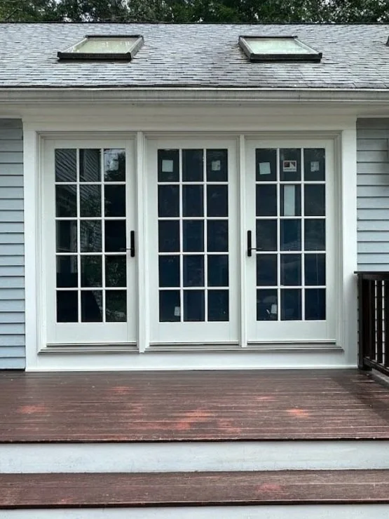 Double doors with glass panes leading to a wooden deck, part of a light blue house with skylights in the roof.