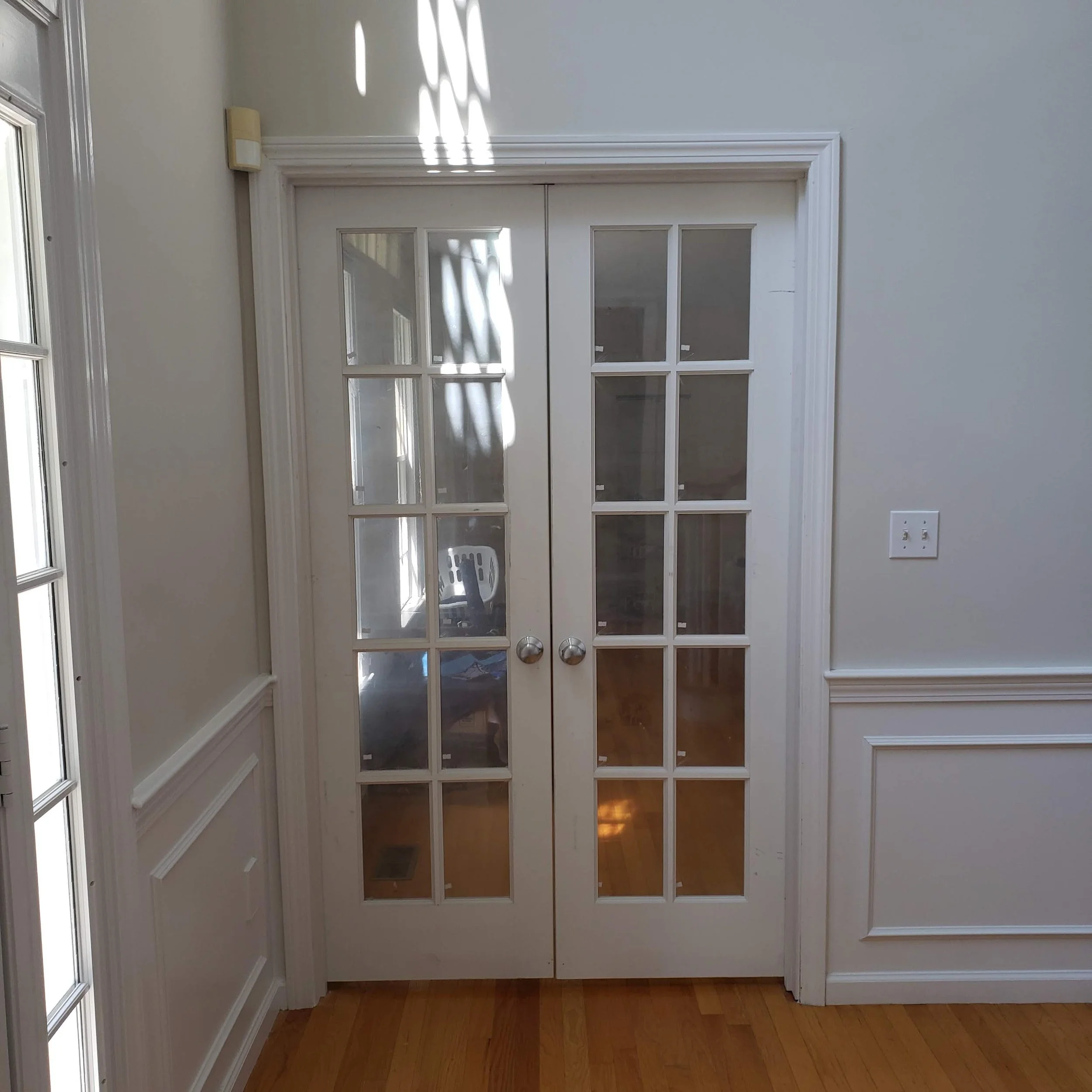 A set of white double doors with glass panes in a home interior. Sunlight creates shadow patterns on the doors and wall. The walls are painted white with decorative trim, and there is a wooden floor.