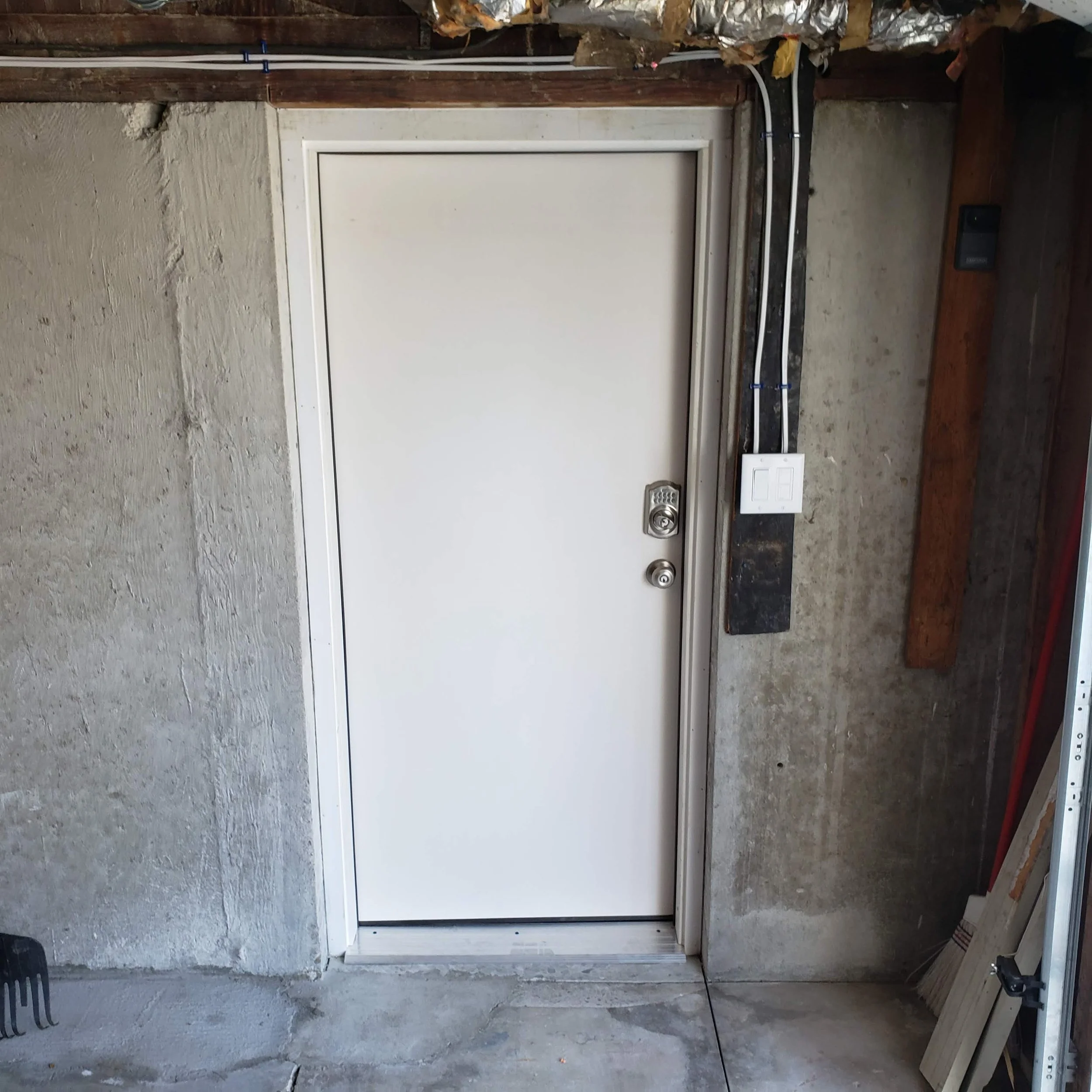 Unfinished interior wall with an electrical outlet, electrical wiring, and pipes near a white door frame, with construction supplies around.