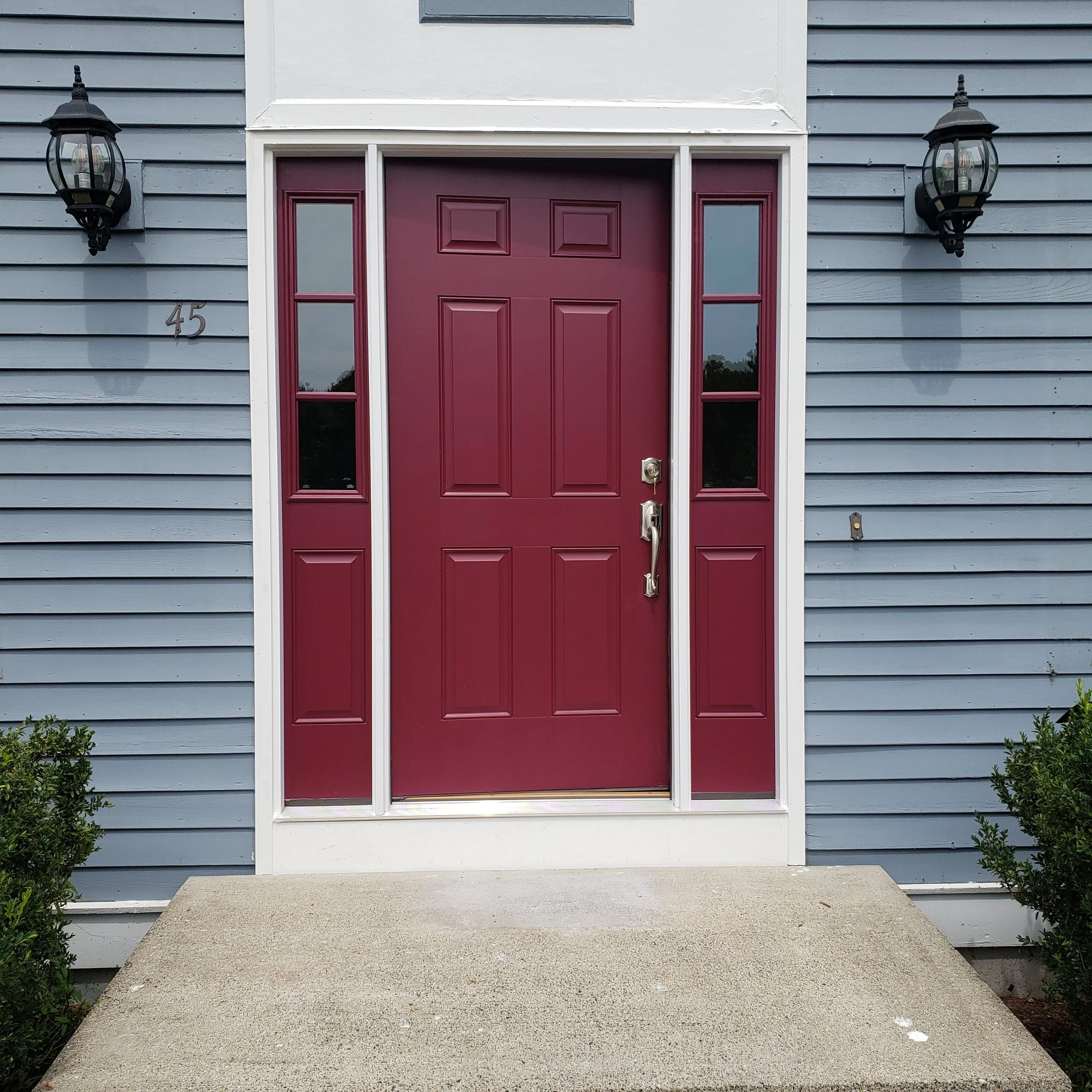 Red front door with six panels, side windows, and a brass doorknob on a house with blue horizontal siding. Two black outdoor wall lanterns flanking the door, house number 45 on the left, small bushes on the sides, and a concrete porch with steps lead