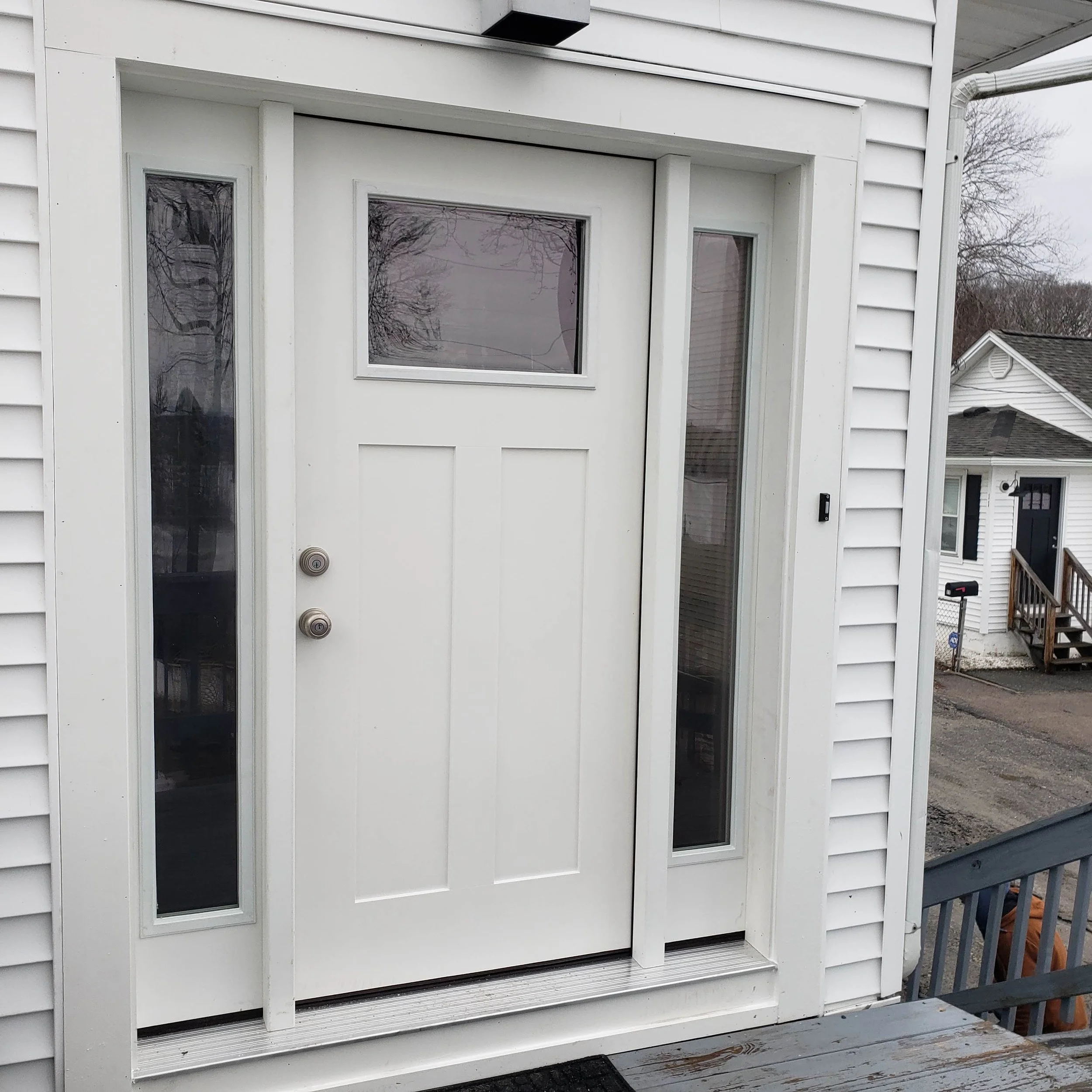 White exterior door with glass window panels and a small window to the right, set in a house with white siding. A black doormat is in front of the door and part of a wooden deck railing is visible on the right.