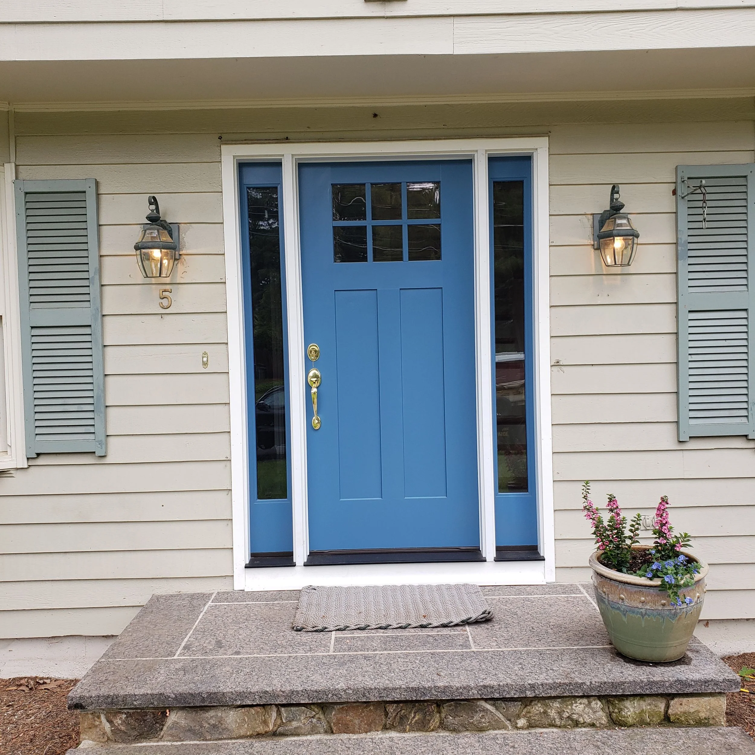 Front door of a house painted blue with glass panels, surrounded by cream siding with light blue shutters, flanked by two wall-mounted lanterns that are lit, with a plant in a large ceramic pot and a doormat in front.