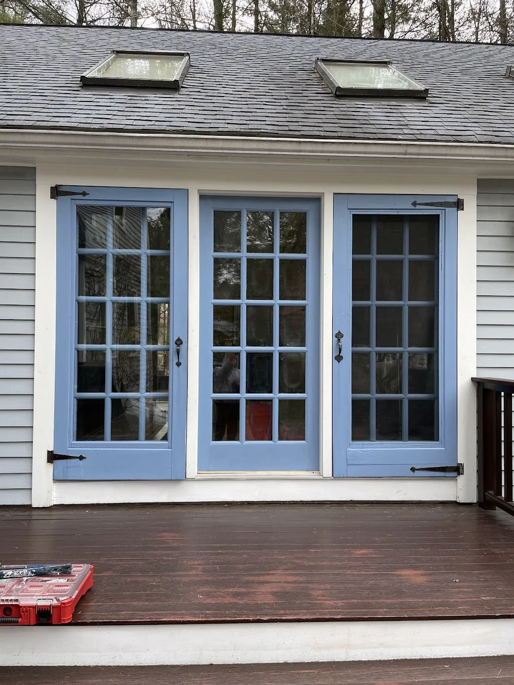 Exterior view of house with blue French doors and shutters on a deck, with a shingle roof and skylights.