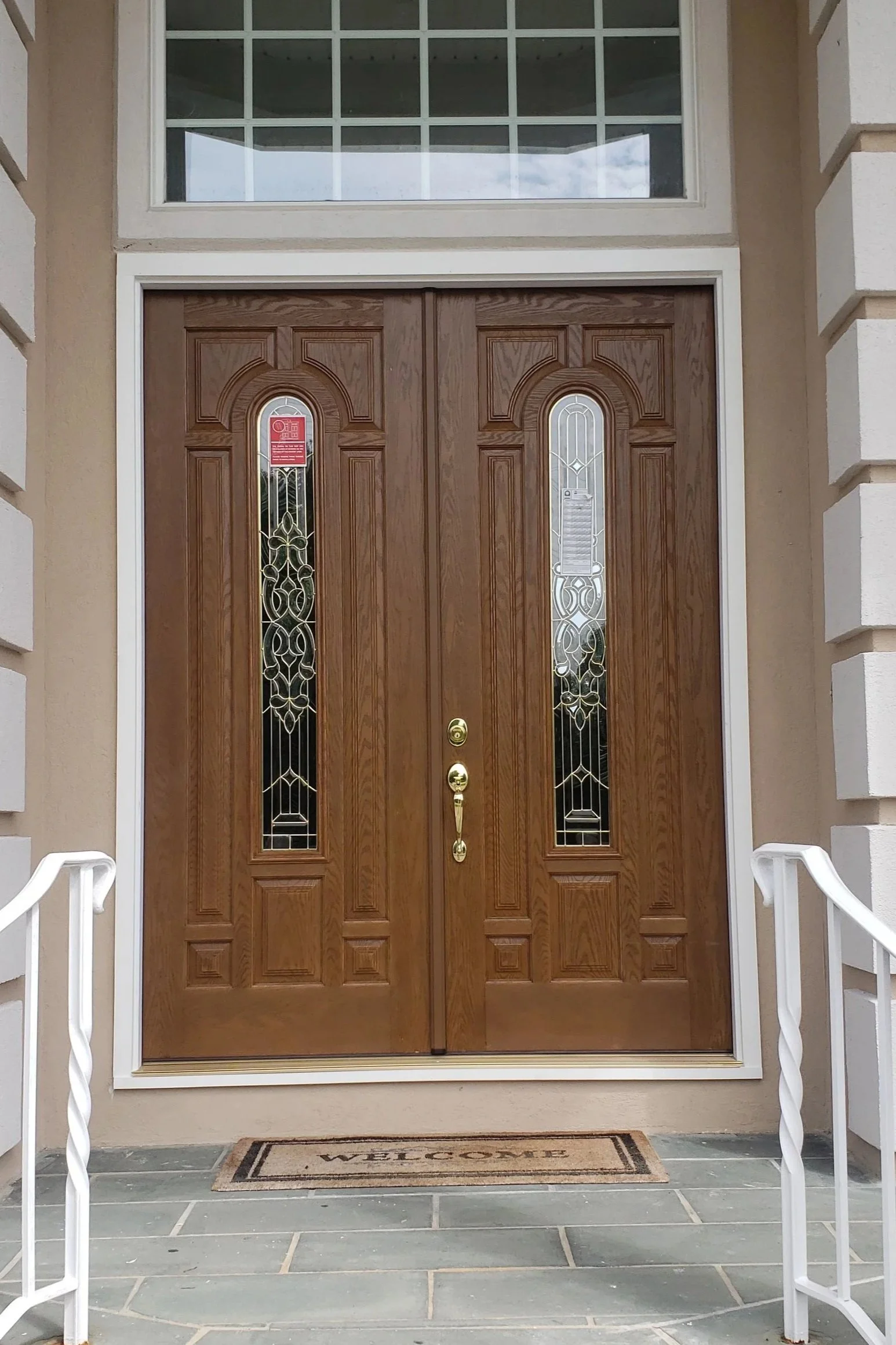 Front door of a house with a welcome mat that reads 'WELCOME'. The door is wooden with glass panels, and there is a white handrail on each side of the steps leading to the door.