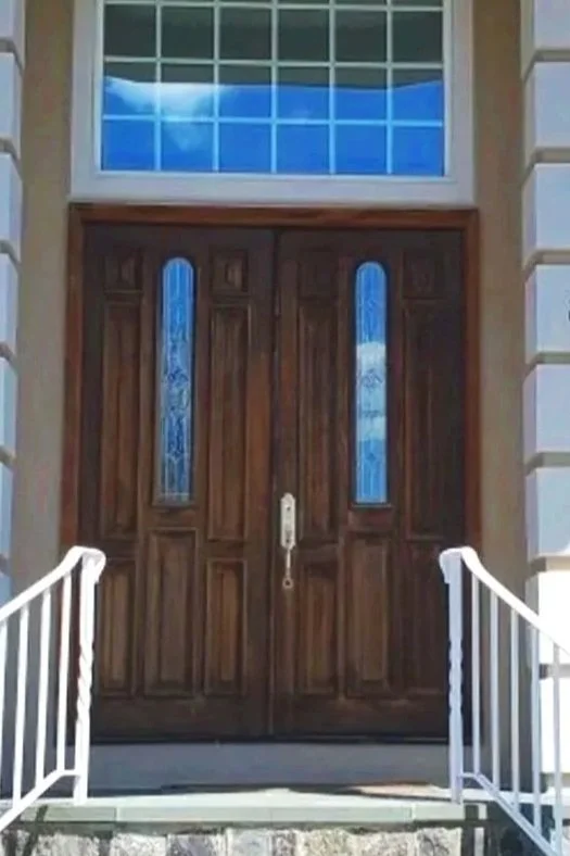 Front door of a house with a wooden double door, glass panels on each door, and a rectangular window above with a view of blue sky and clouds.