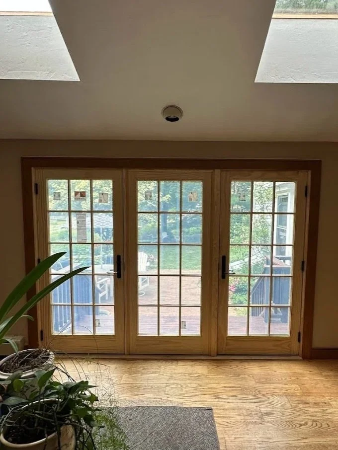 Interior view of a room with a large wooden framed glass sliding door leading to a wooden deck outside, with greenery visible beyond.