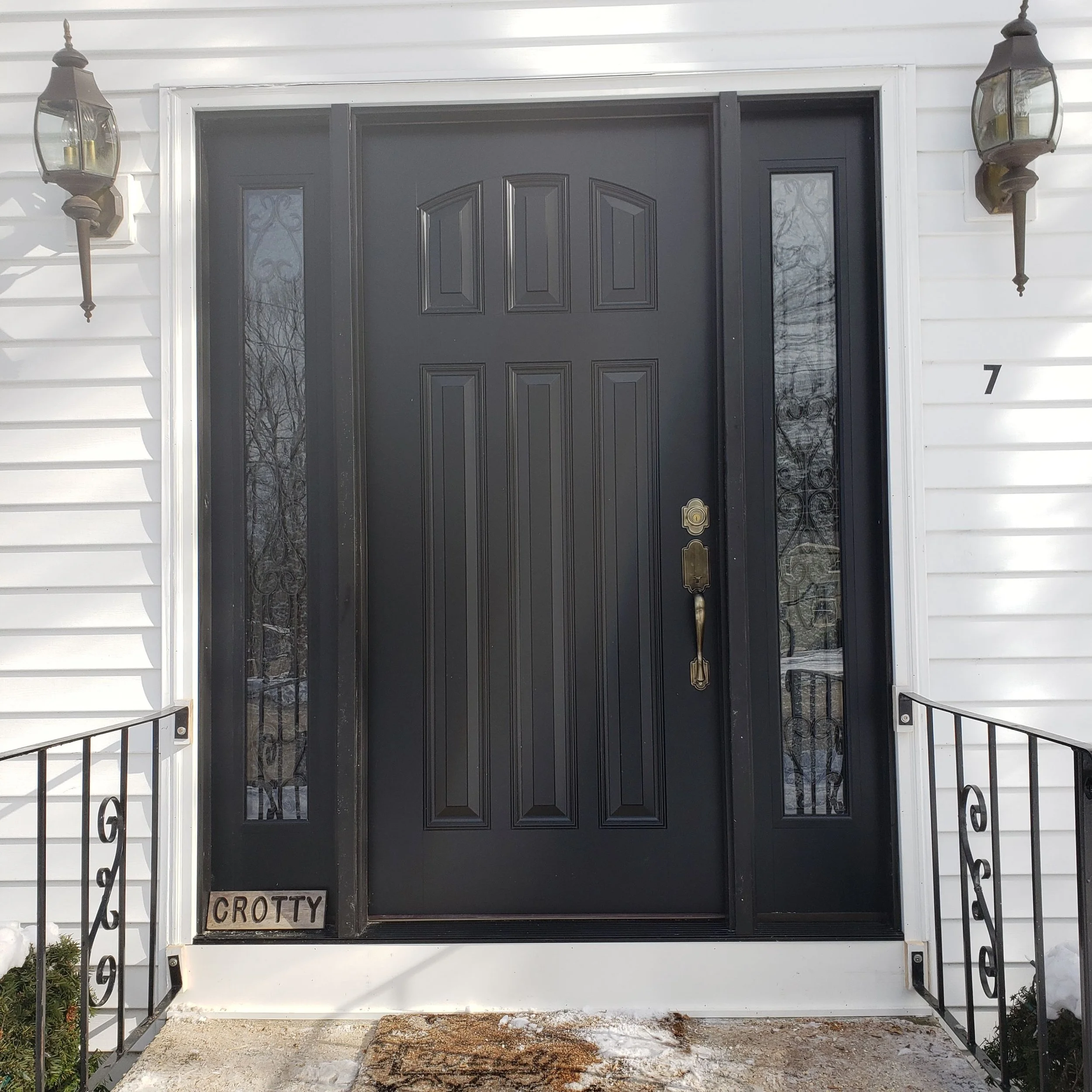Black front door with decorative glass panels on either side, mounted on a white house with white siding and a black metal railing, with a doormat and small plants near the entrance. Two wall-mounted lantern-style light fixtures are on either side of