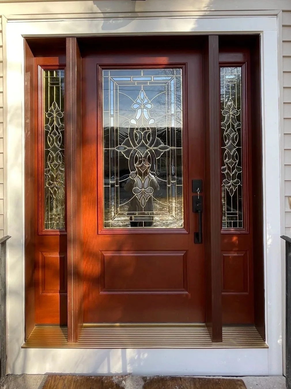 Wooden front door with decorative glass panels and sidelights, surrounded by a white frame, with a small metal grate at the threshold.