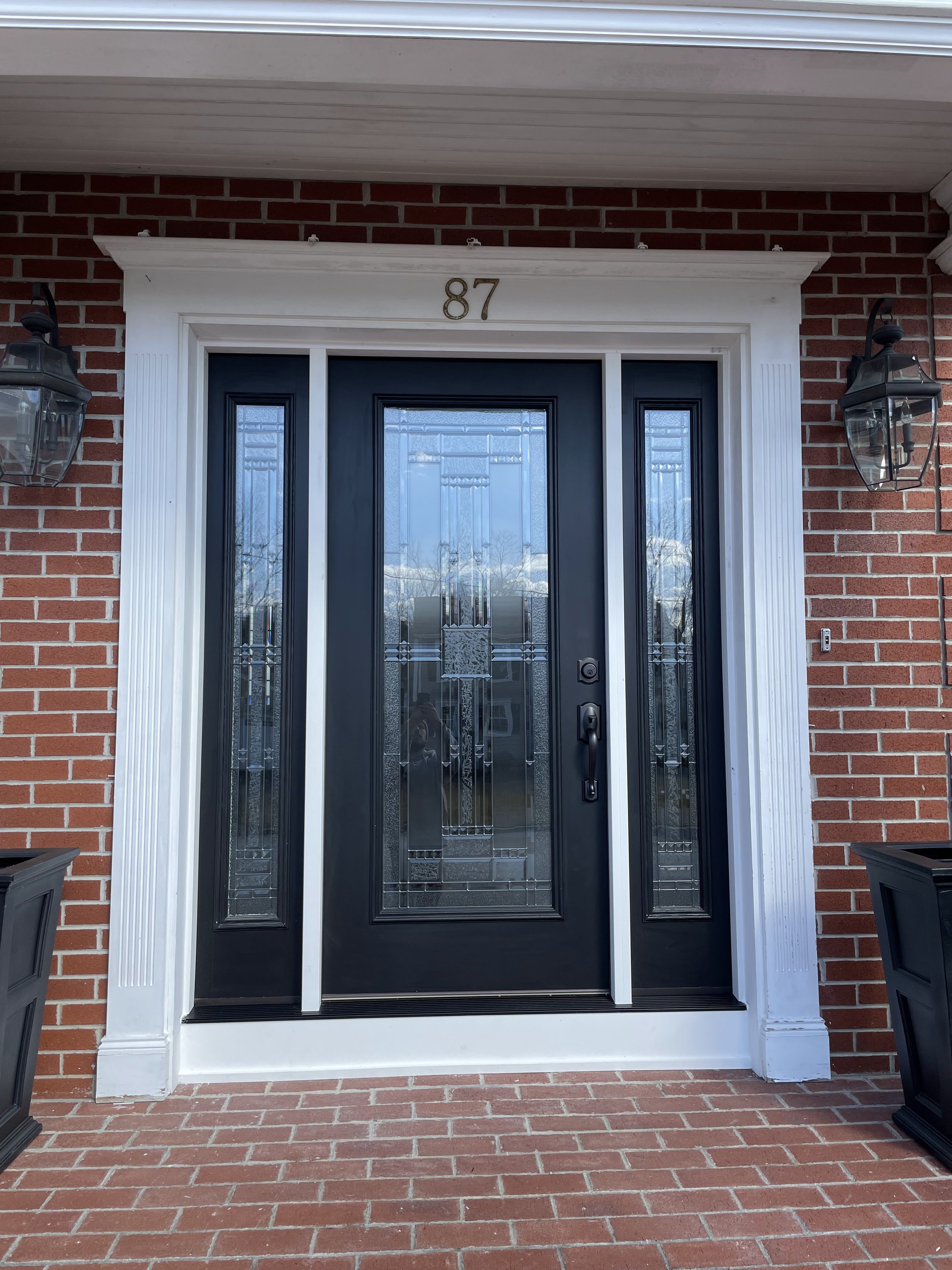 Front door with black frame, glass panels, and house number 87 above the door, flanked by black outdoor lanterns mounted on red brick wall.