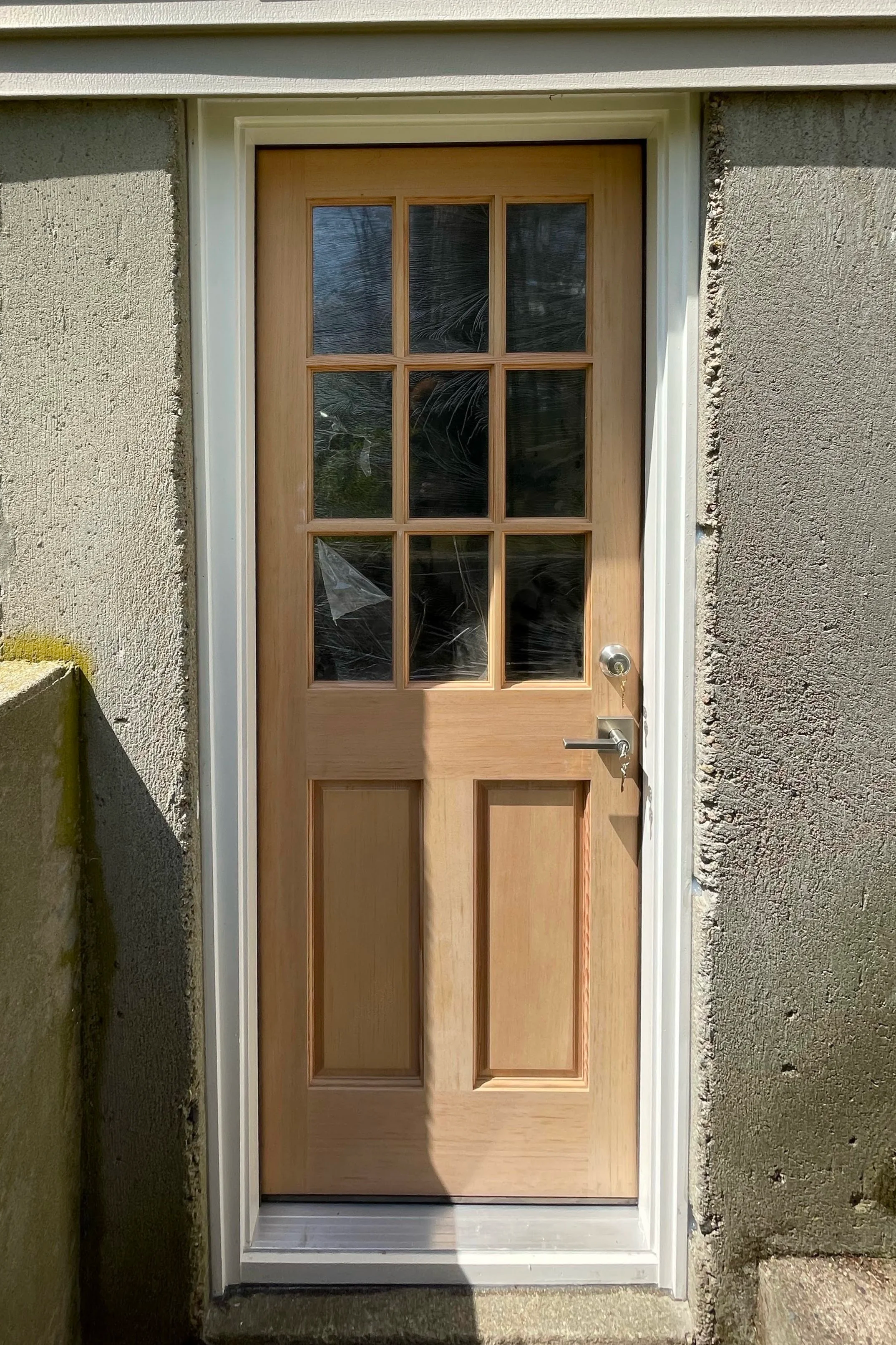 Front door with a light-colored wooden finish, featuring nine glass panels arranged in a grid pattern, with a silver door handle and lock, set in a doorstep with concrete and rough-textured exterior walls.