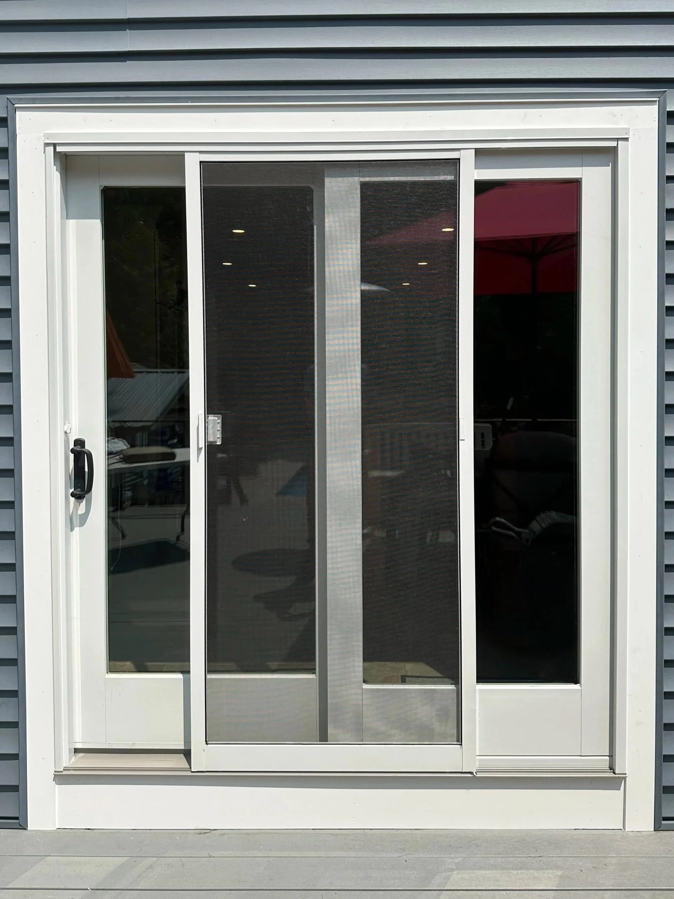A modern exterior sliding glass door with white frame, black mesh screen, and a silver handle, set against a grey building with horizontal siding.