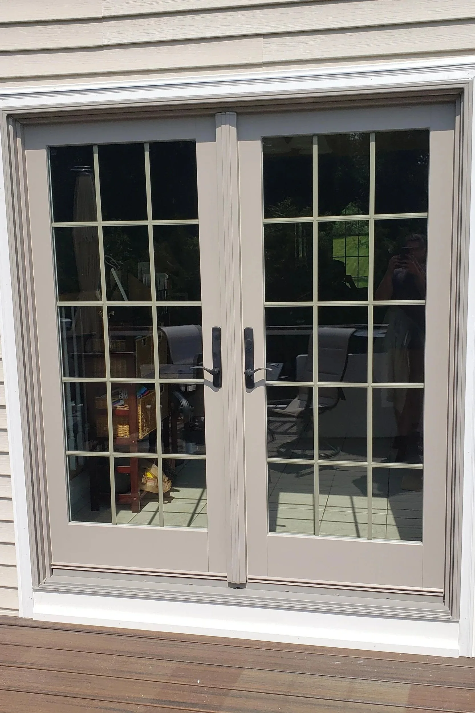 View of a white sliding glass door with multiple panes, opening to a deck with wood planks. Inside, there are chairs, a table, and various household items visible through the glass.