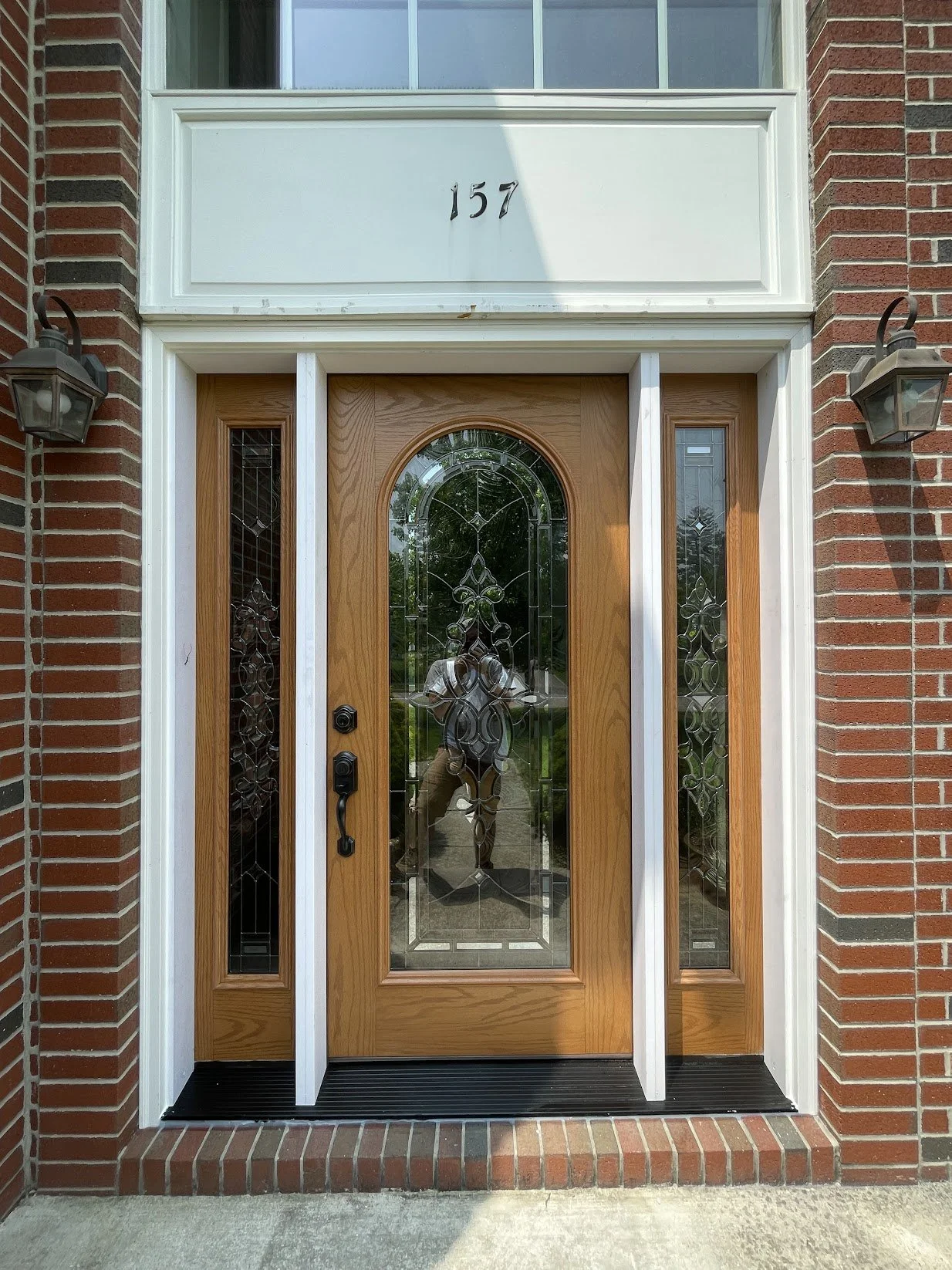 Front door with a wooden frame, decorative glass panel, and side glass panels, set in a brick wall with house number 157 above. Two outdoor wall-mounted lanterns flank the door.