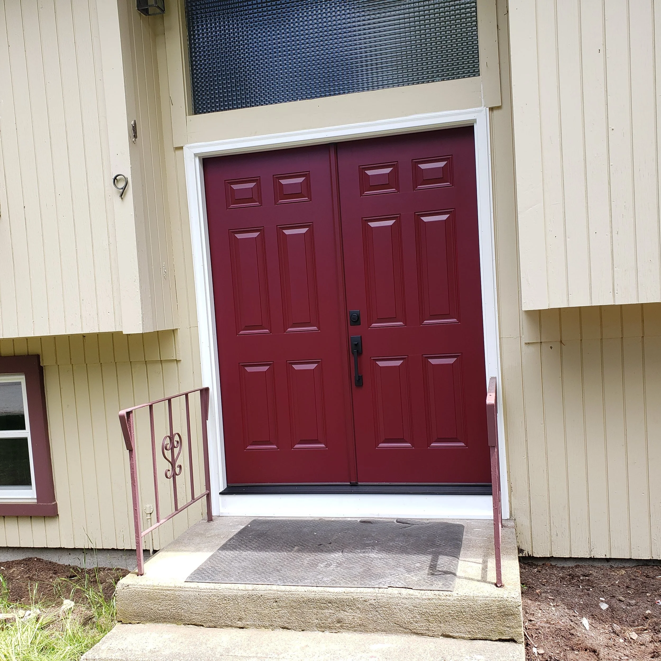 Front entrance of a house with a red garage door, white framing, stairs and handrails, beige siding, and a welcome mat on the porch.
