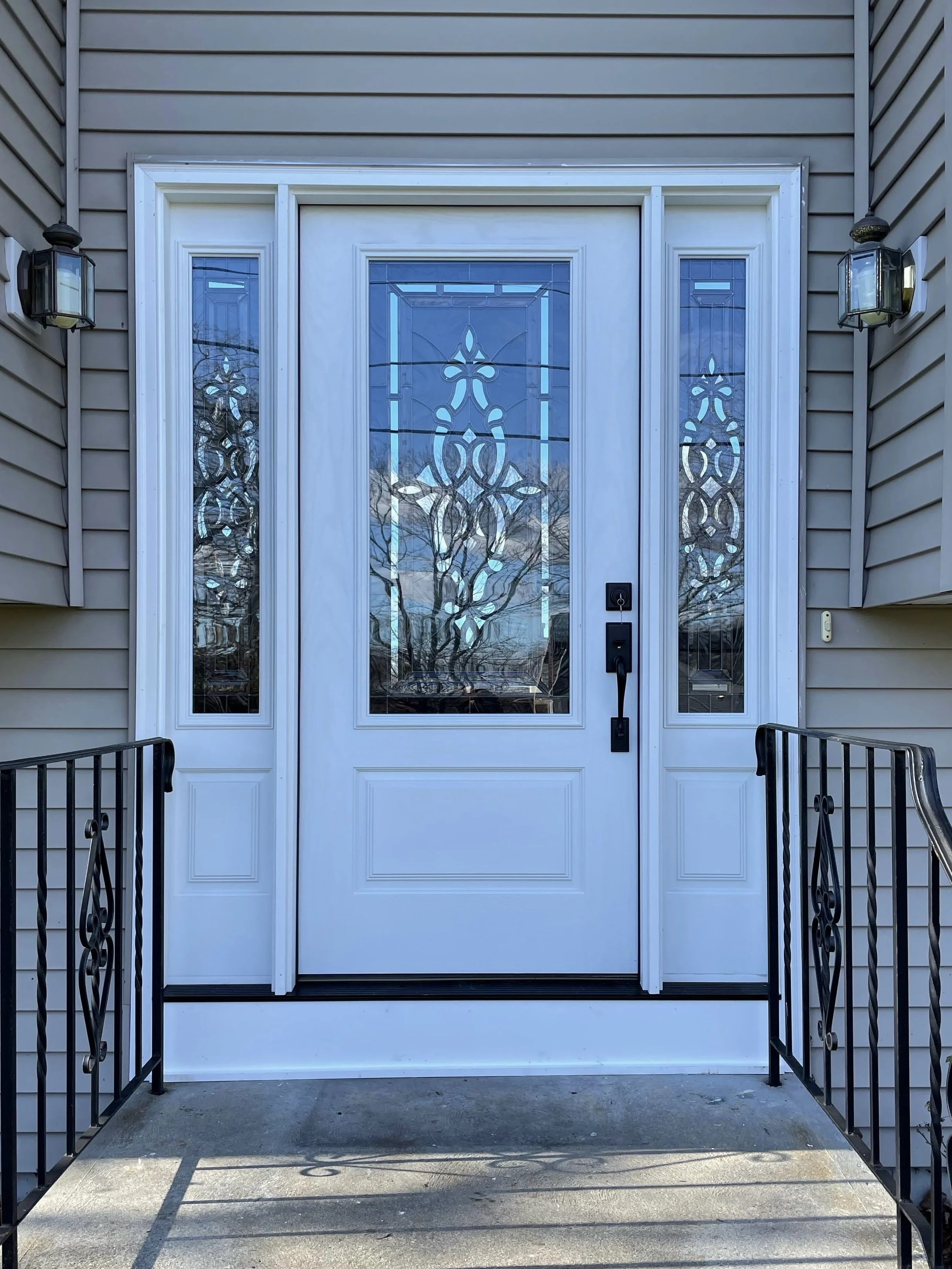 Front door with glass panels, flanked by side windows, with black metal railings on both sides, and an exterior light fixture on each side of the house.