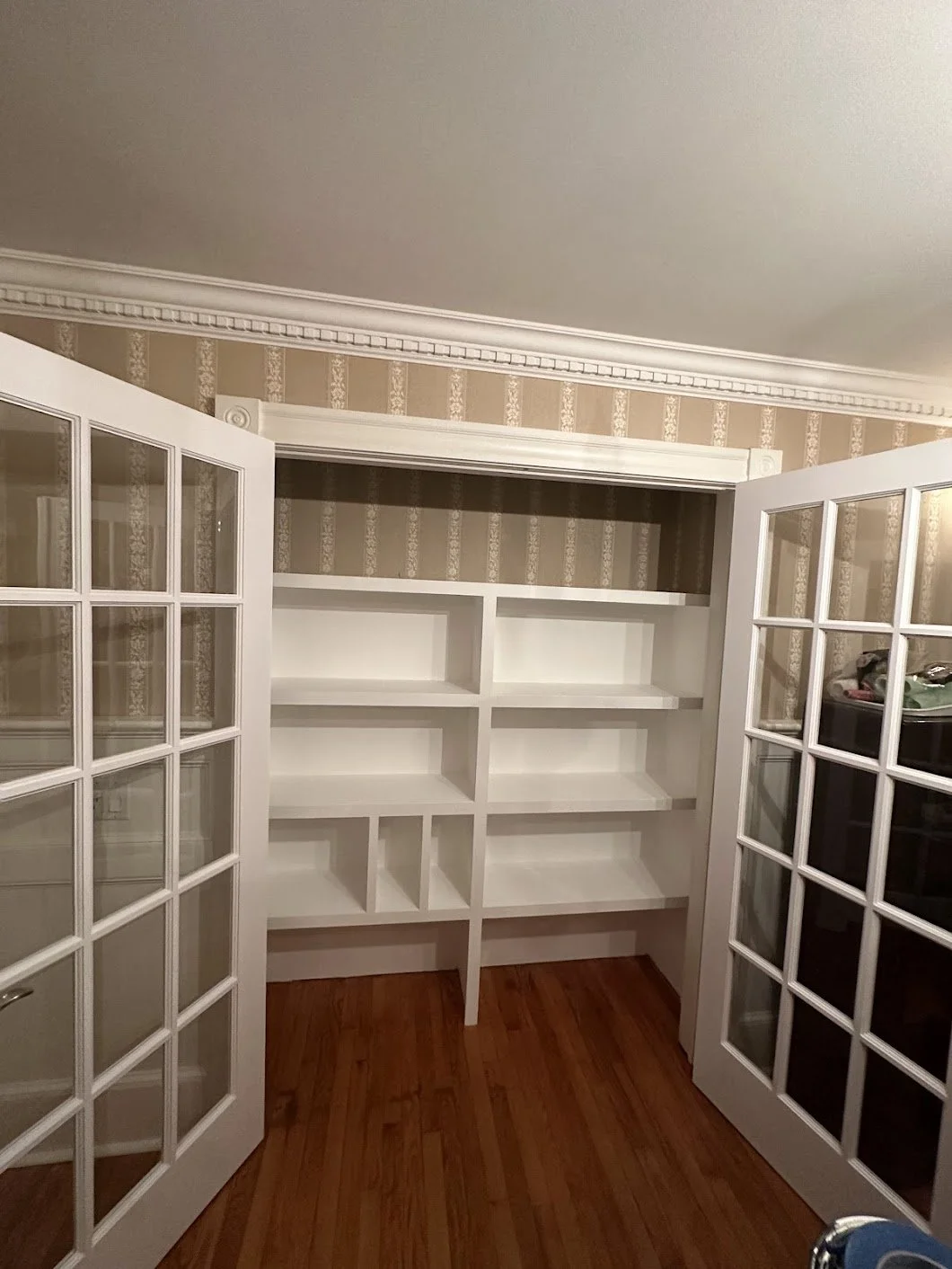 White built-in bookshelves with glass-paneled French doors in each opening, set against beige and gold wallpapered wall and hardwood floor.