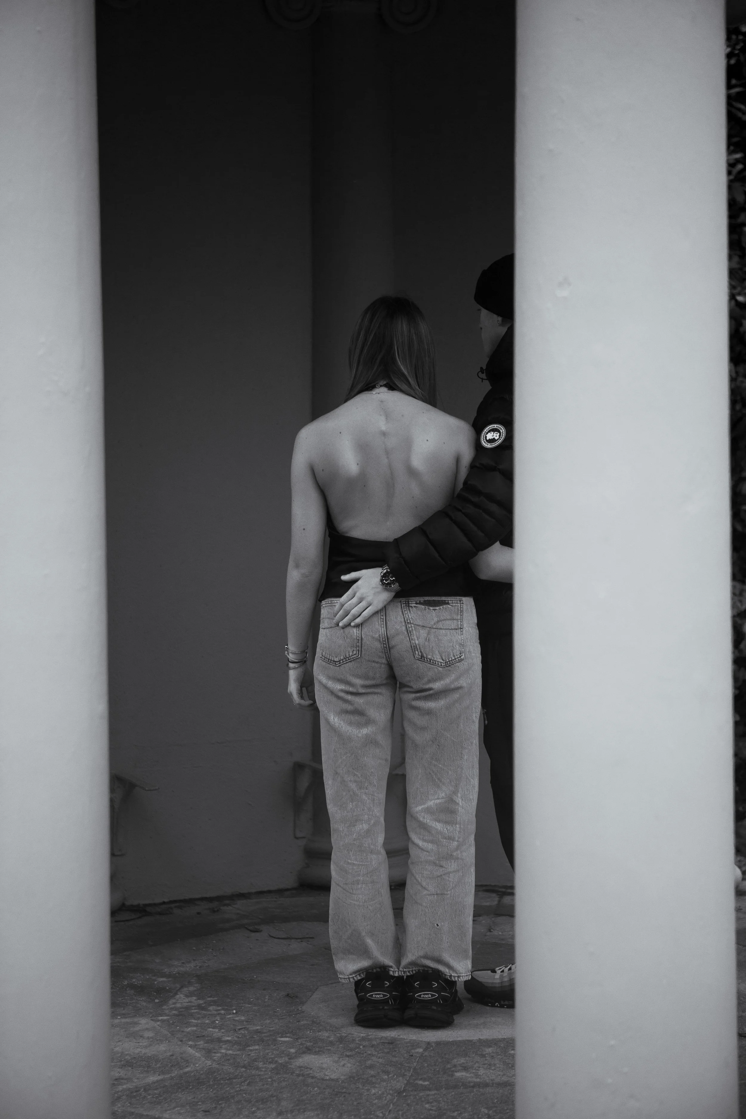 Black and white portrait photograph of a young woman with scoliosis, shown from behind, standing between architectural columns while being supported by another figure, exploring themes of care, vulnerability, and the human body.
