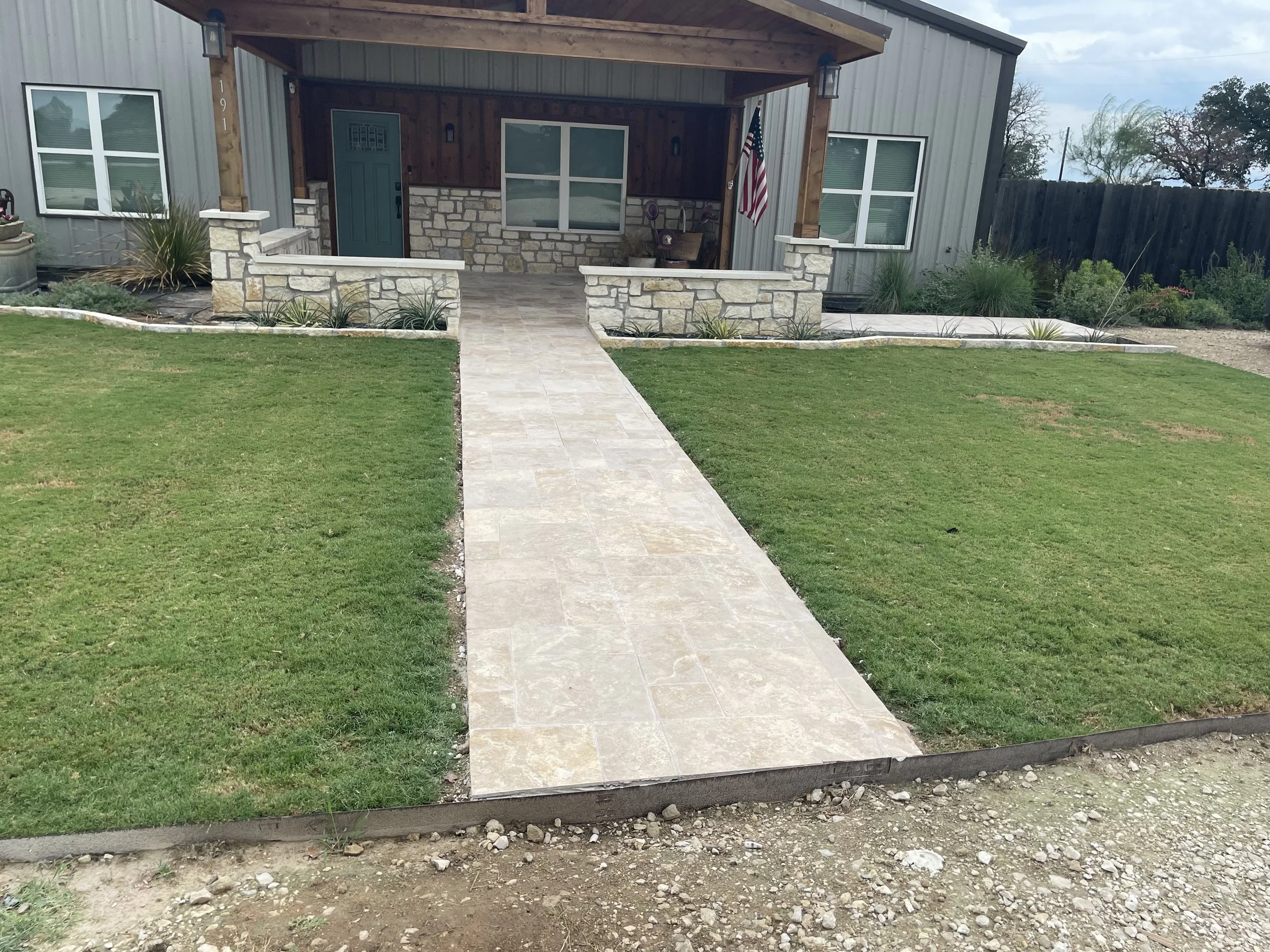 Front view of a house with a concrete pathway leading to the front porch, which has a stone and wood facade, green door, and American flag. The front yard has well-maintained grass and garden beds.