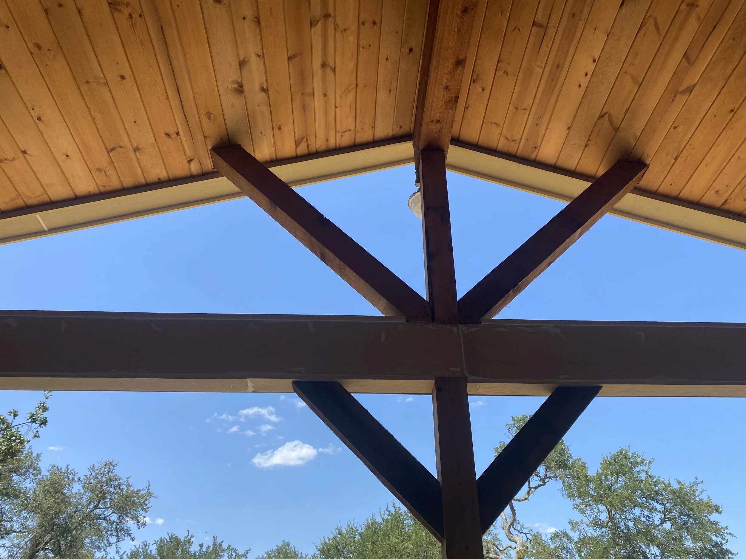 Looking up at the underside of a wooden pergola roof with support beams against a blue sky, with some trees visible at the bottom and sides.