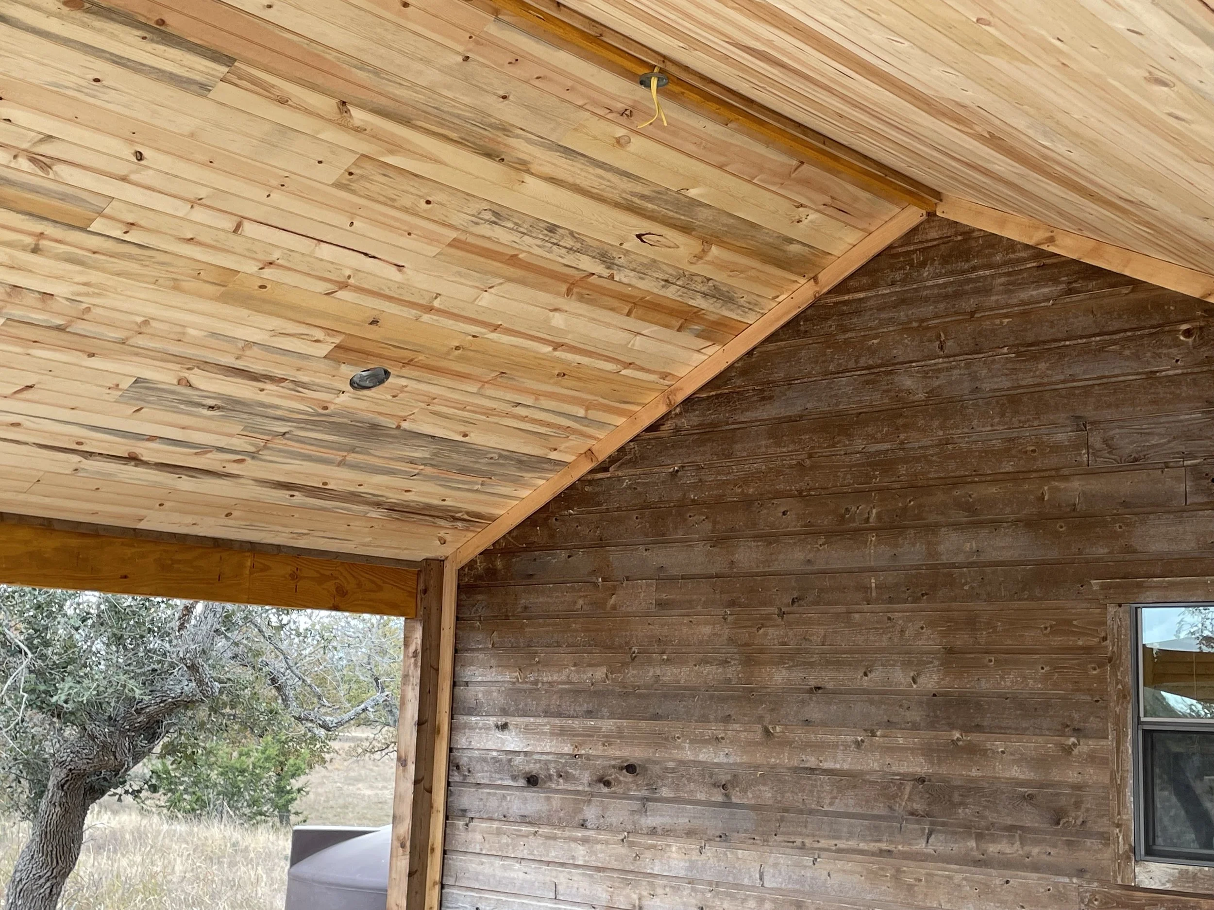 Photo of a wooden ceiling with light-colored wooden planks and a darker wooden wall below. Part of a window with a tree outside is visible in the lower right corner.