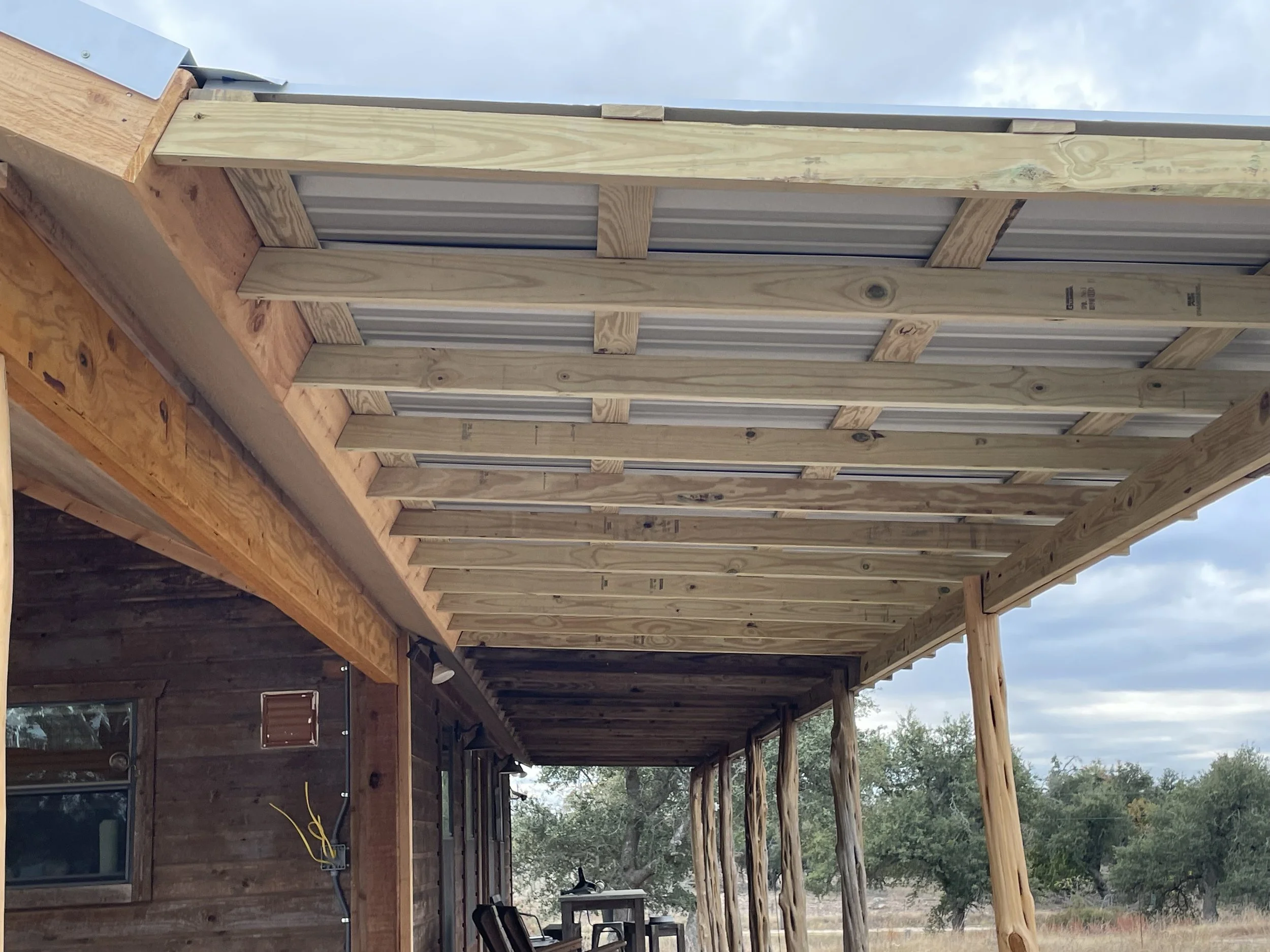 Under-construction outdoor patio with wooden framing and metal roofing, attached to a house with a window and electrical wiring visible.