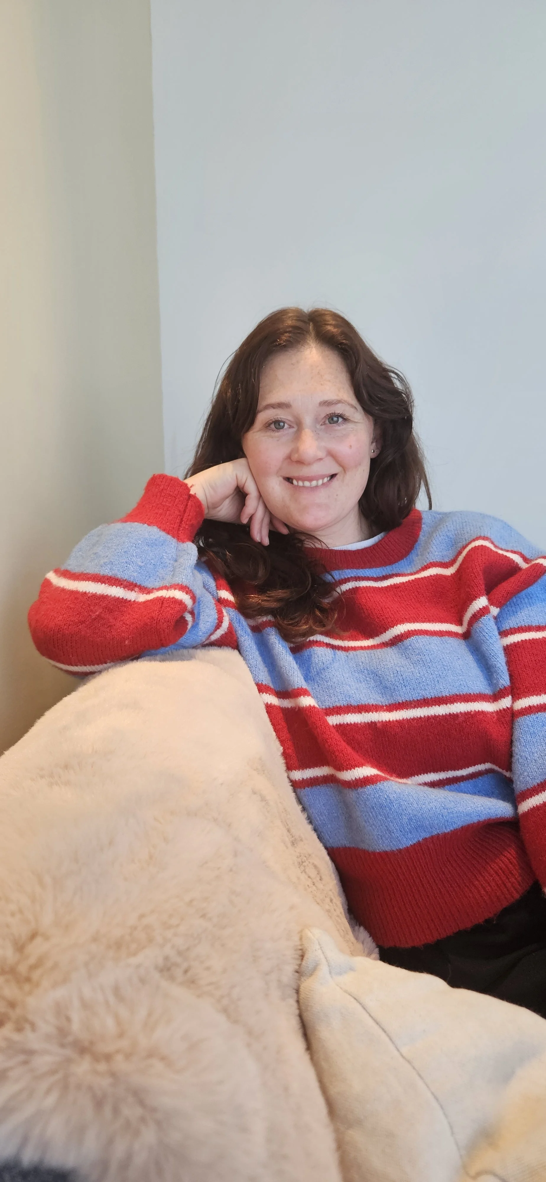 A woman with long, wavy brown hair and freckles, wearing a colorful striped sweater, sitting relaxed with her hand resting on her face and smiling at the camera