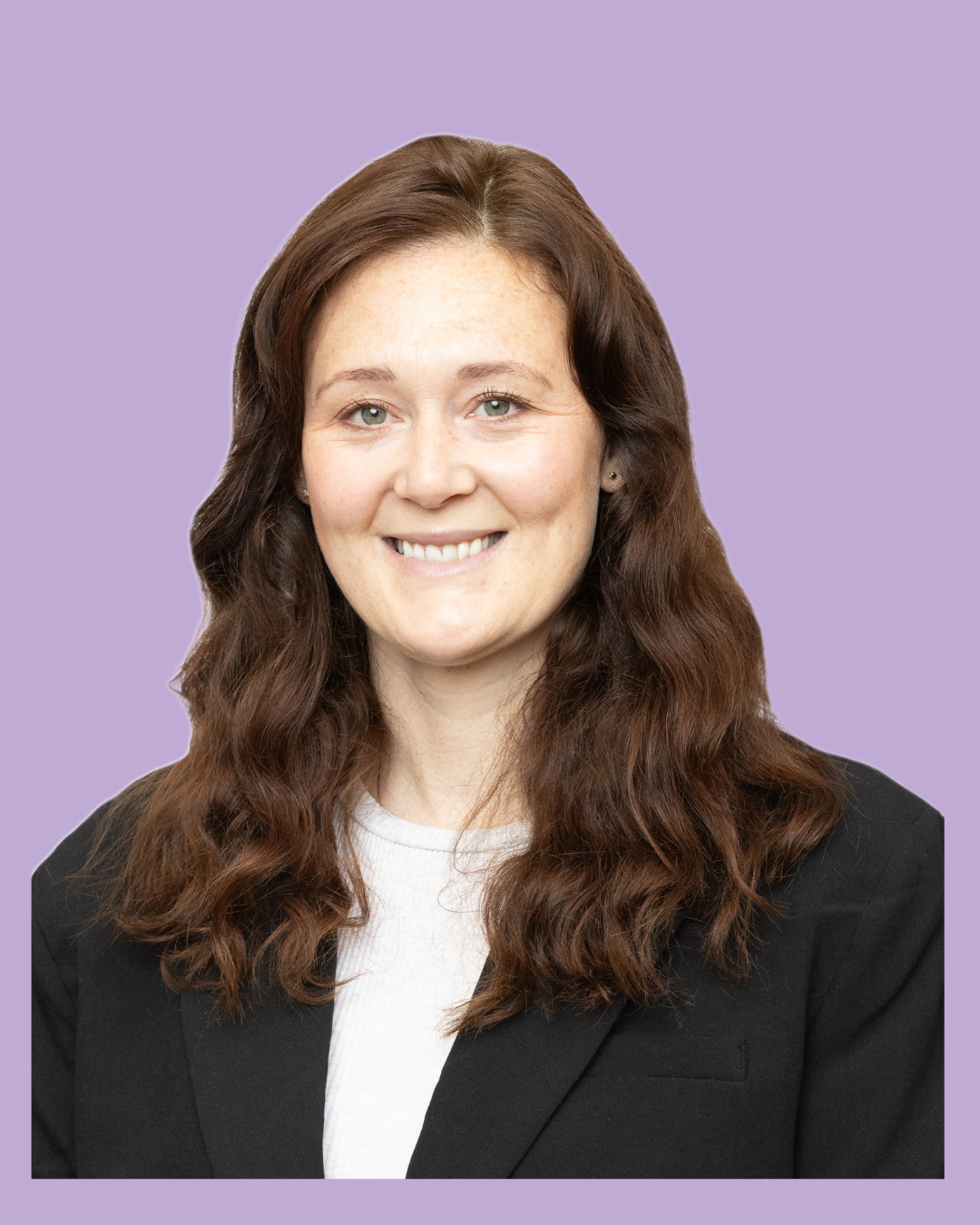 A woman with long, wavy brown hair and light skin, smiling, wearing a black blazer over a white top, with a lavender background.
