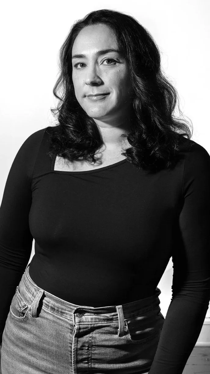 Black and white portrait of a woman with wavy dark hair, wearing a black long-sleeve top and jeans, standing against a light background with a slight smile.