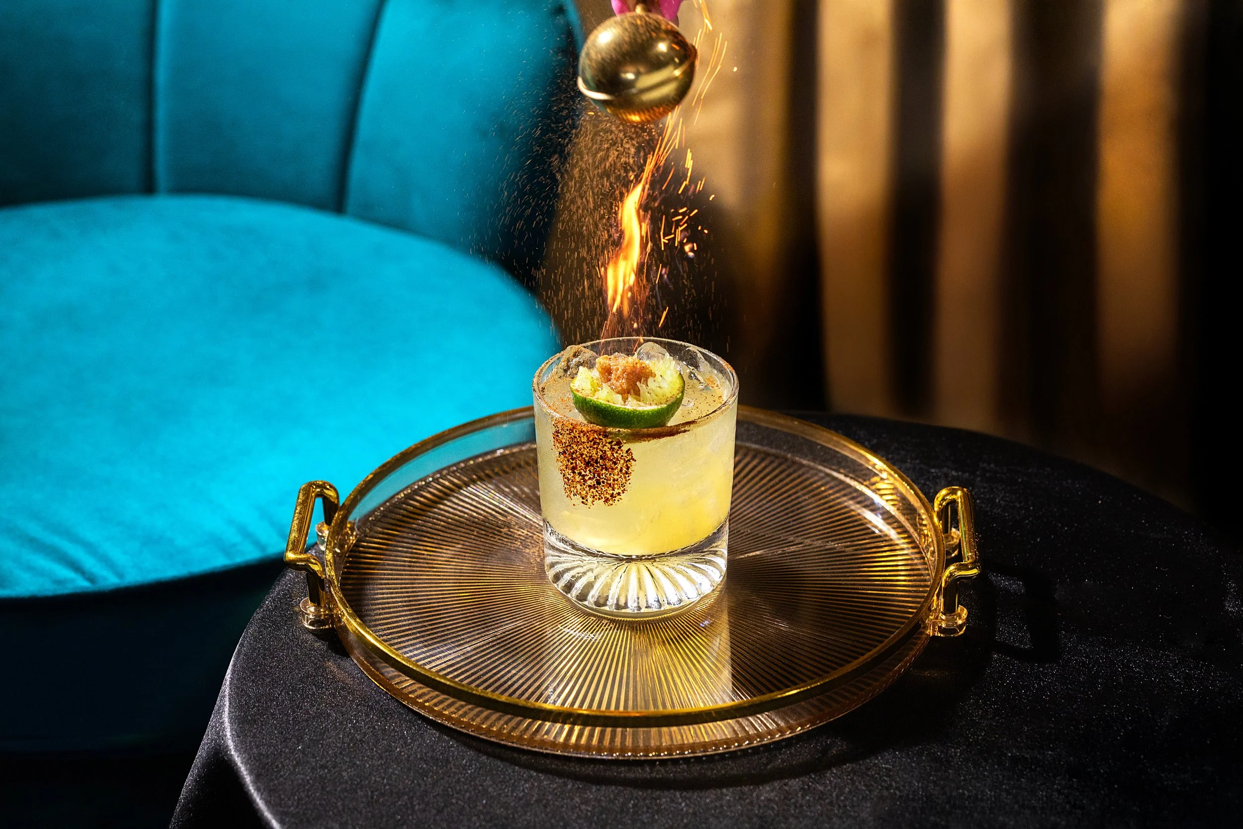 A cocktail drink garnished with lime, topped with a sparkler firework, served on a golden tray, with a blue upholstered chair in the background.
