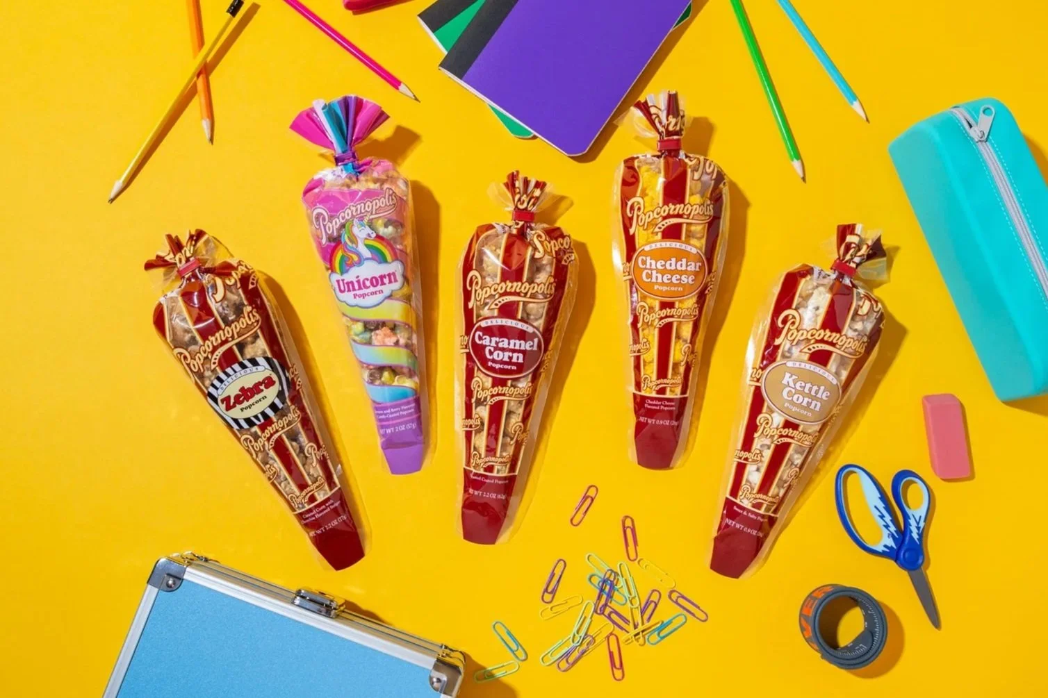 Five popcorn cones with colorful branding and flavors arranged on a yellow background. Surrounding items include a blue notebook, pink eraser, blue scissors, a roll of black tape, a blue pencil case, colorful paper clips, a purple book, and two yellow and pink pencils.
