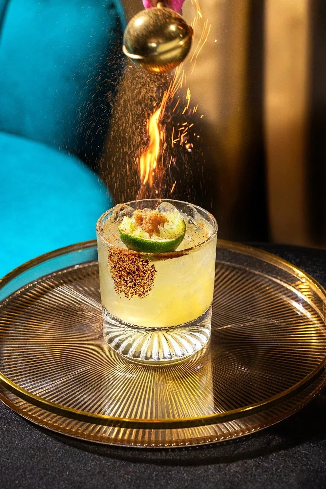A bartender flames a cocktail garnished with lime, chili powder on the rim, and a lime slice with chili topping, placed on a gold tray.