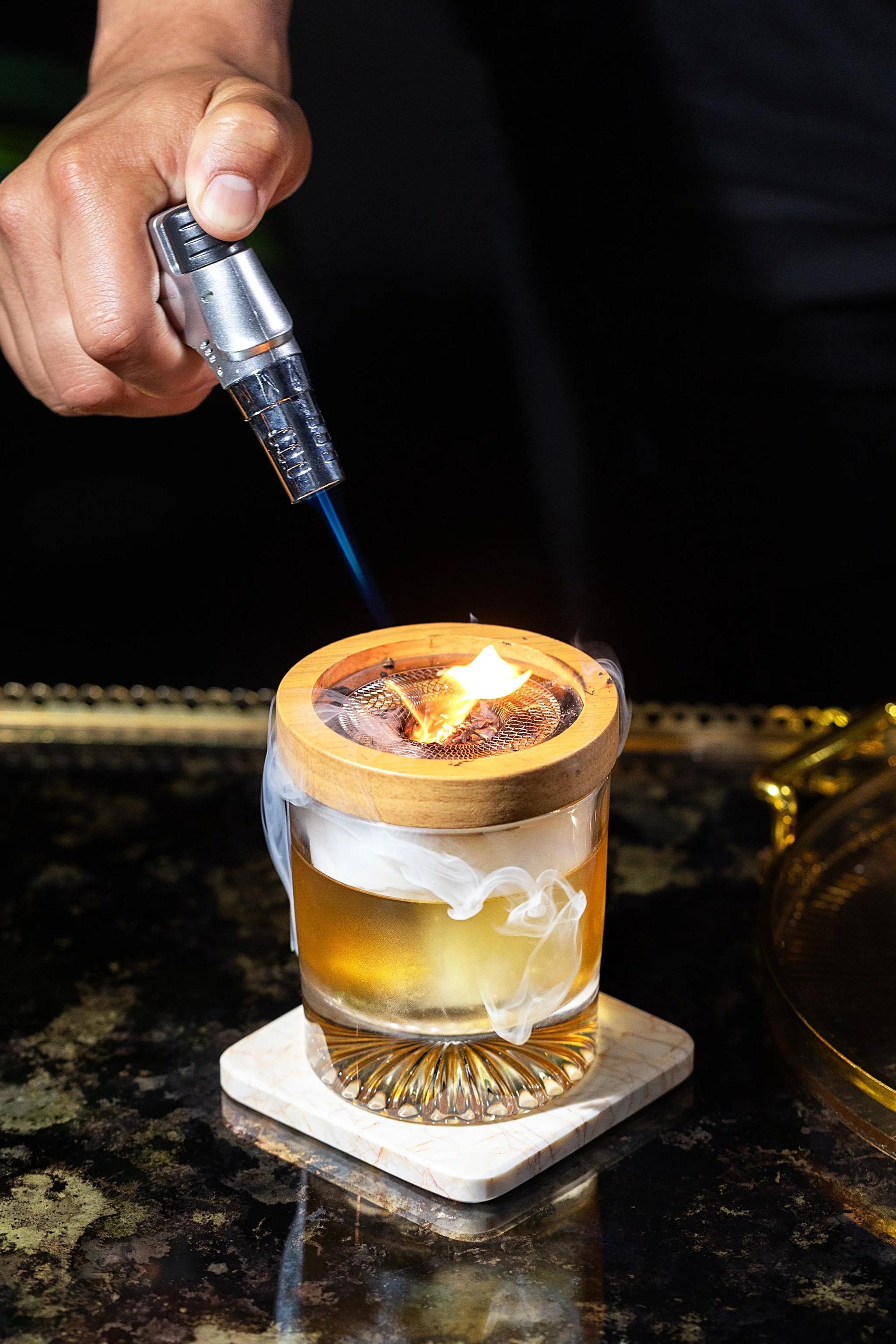 A person holding a small metal torch with a blue flame over a lit candle in a glass container. The candle is emitting smoke and is placed on a square marble coaster.