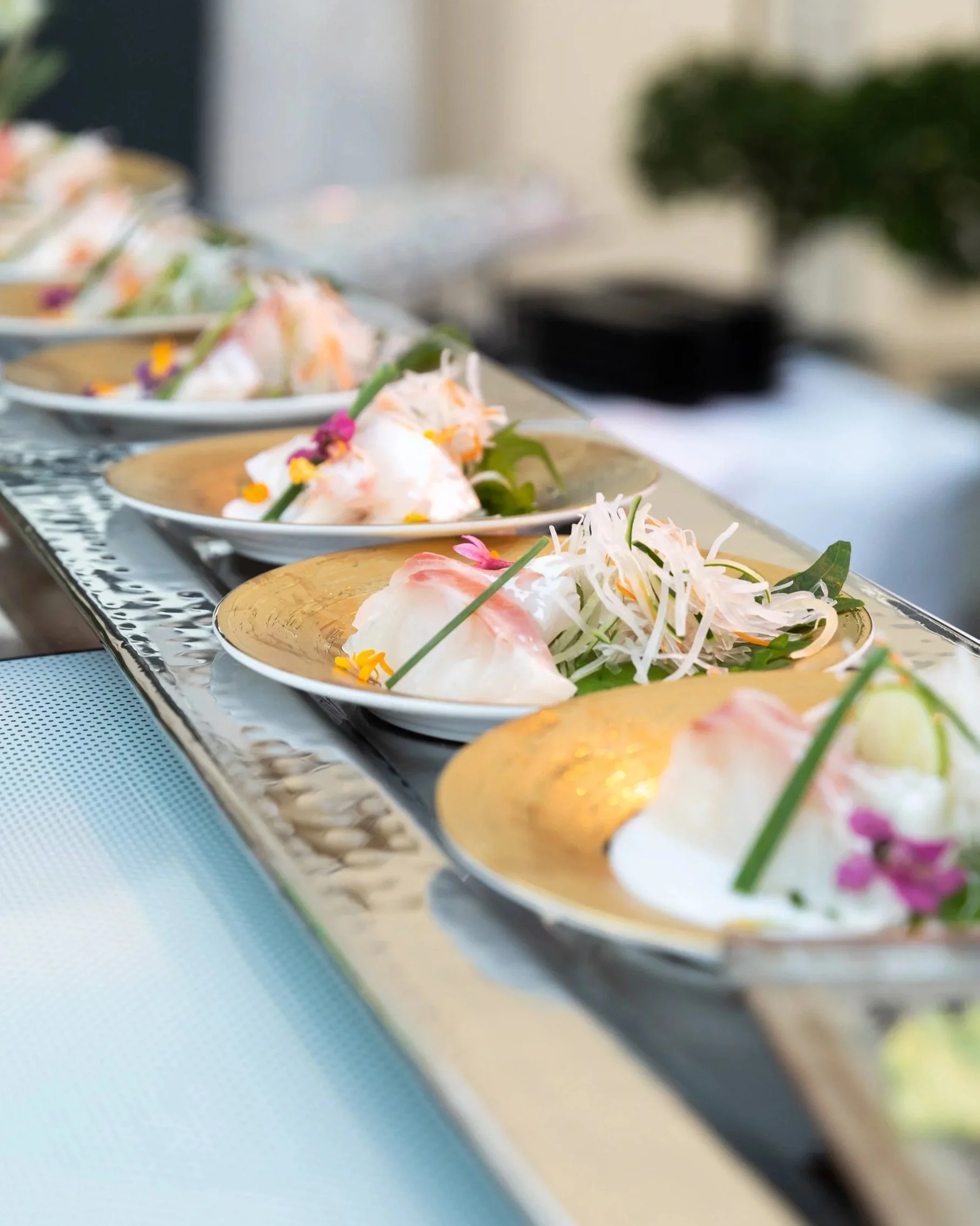 Close-up of multiple plates with steamed pink and white dumplings garnished with edible flowers and greens on a silver tray.