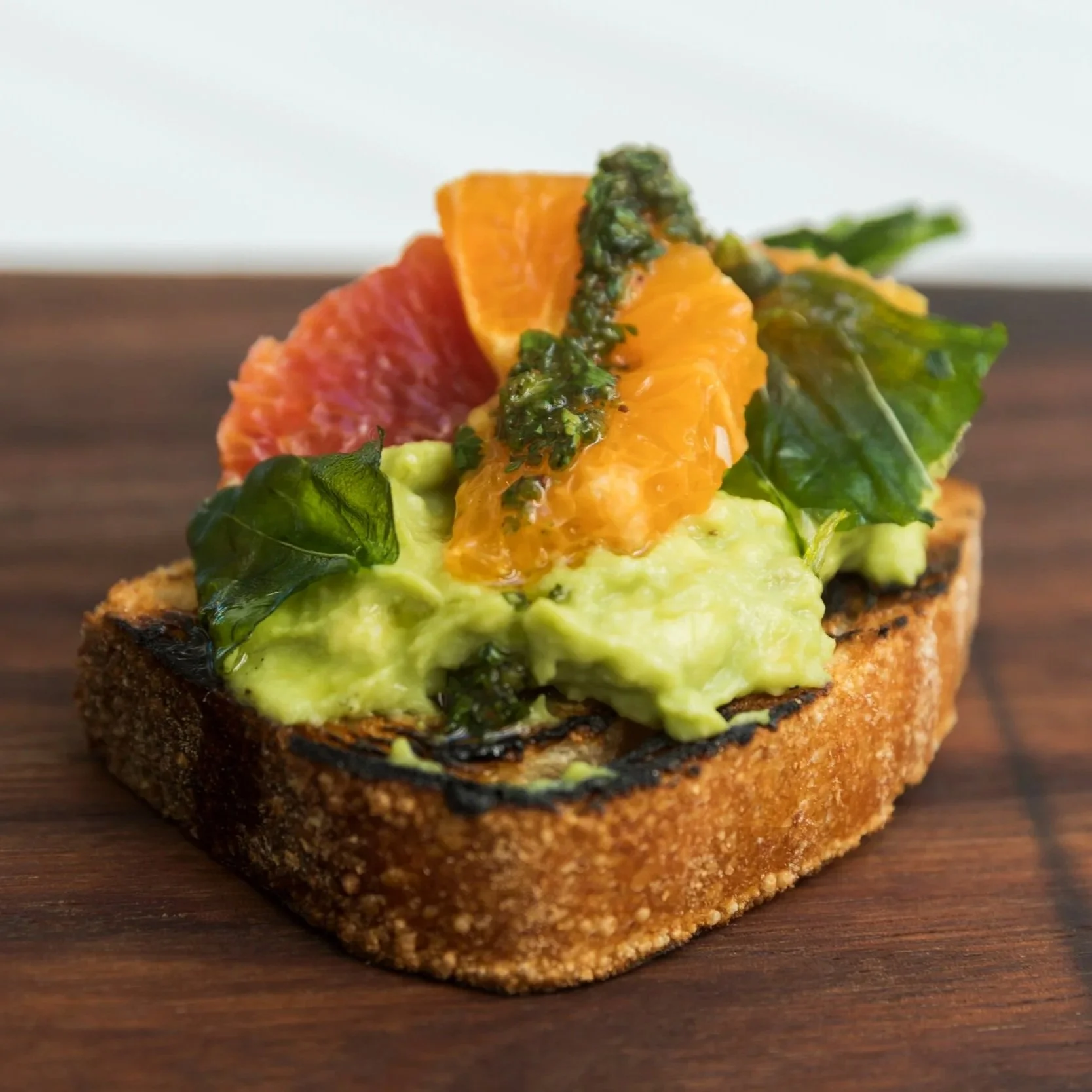 Close-up of a toasted bread slice topped with guacamole, a slice of grapefruit, a slice of orange, greens, and a drizzle of green sauce.