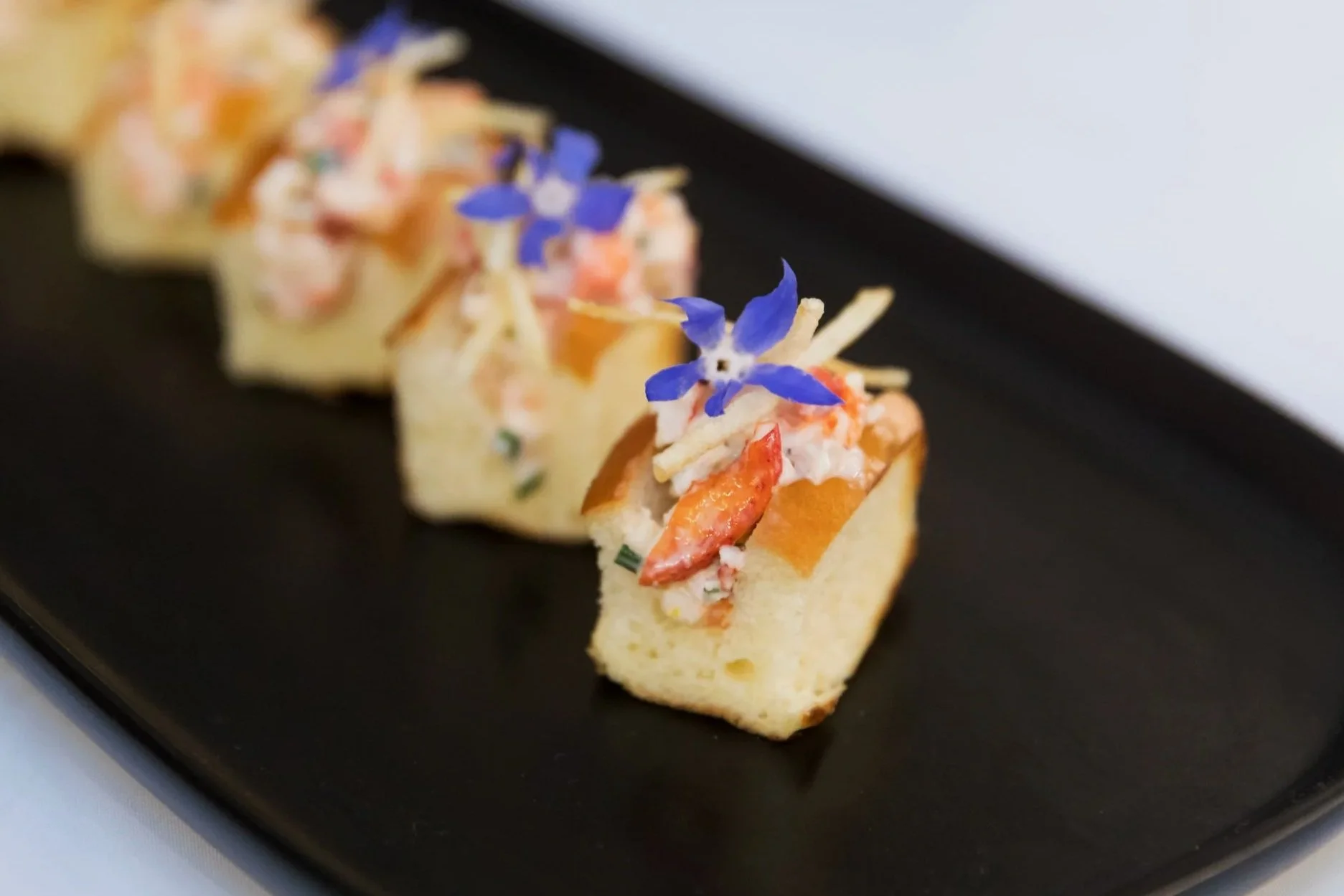 Close-up of an appetizer with seafood, herbs, and edible flowers on a black rectangular plate.