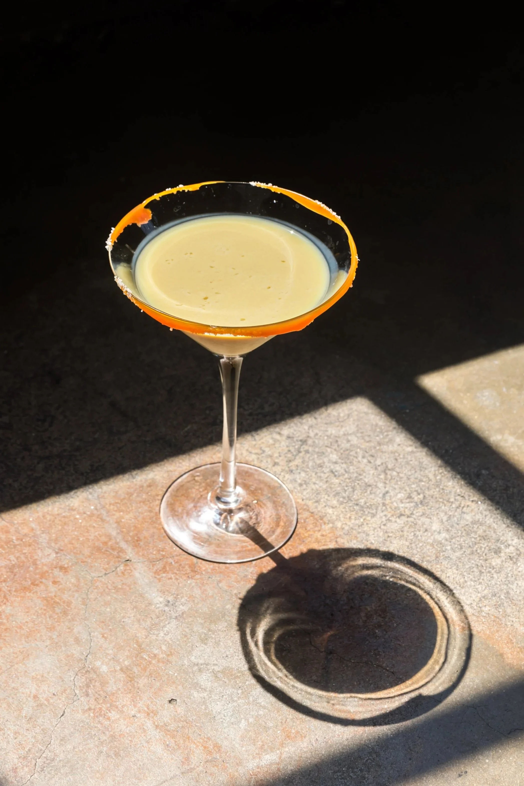 A martini glass with a salted rim, filled with a yellow cocktail, placed on a concrete surface with shadows.