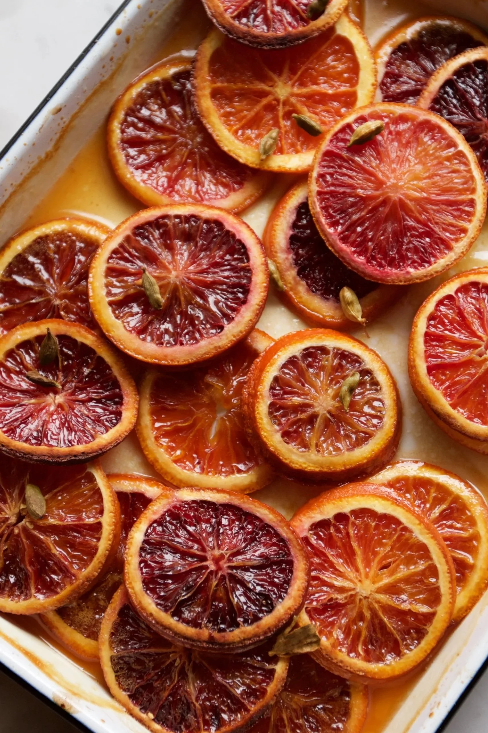 A close-up view of a dish with slices of blood oranges arranged in a baking dish, some garnished with green cardamom pods.