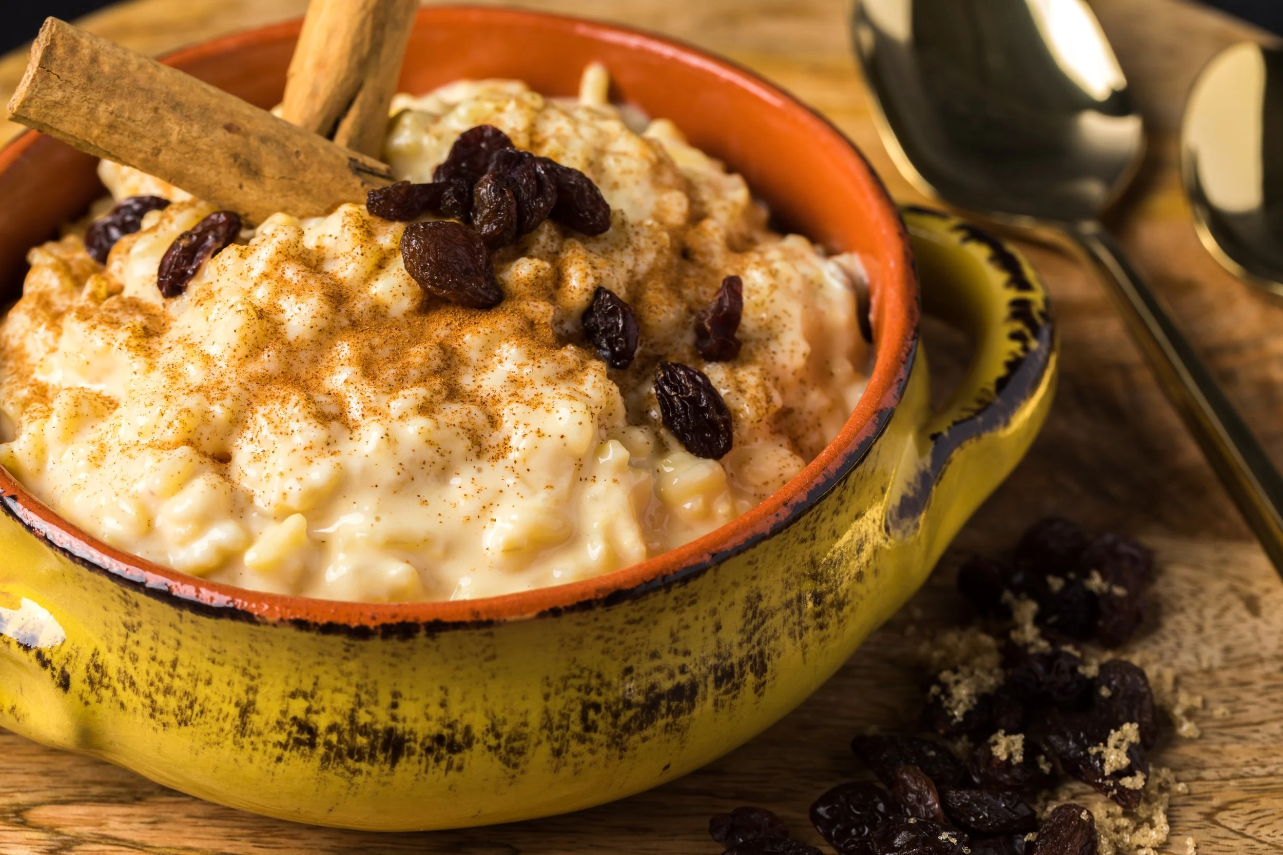 Baked rice pudding topped with raisins, cinnamon, and cinnamon sticks in a yellow ceramic bowl on a wooden surface.